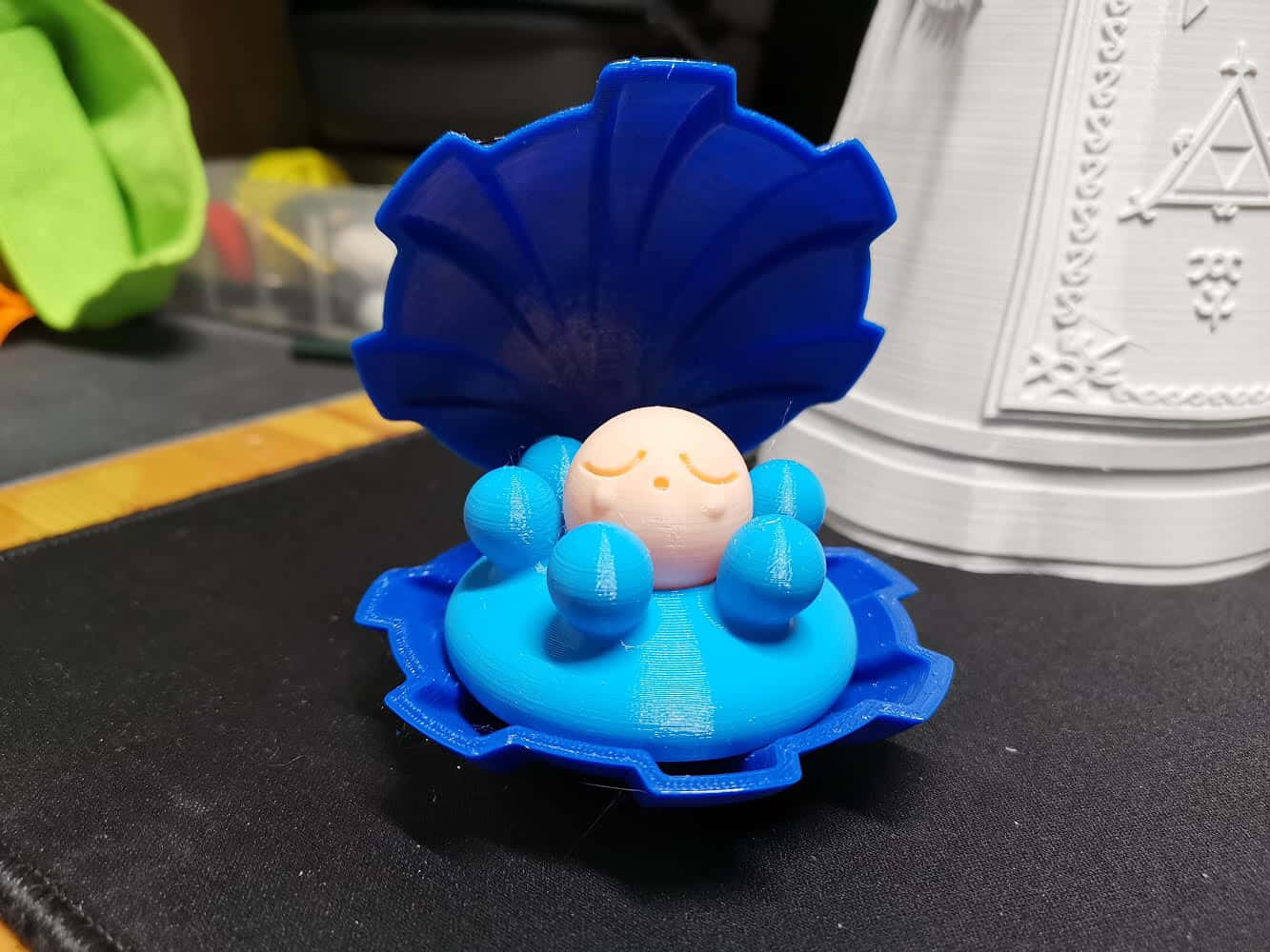藍色 桌布 壁紙 — 一個3D列印的藍色貝殼玩具，裡面有一顆微笑的珍珠，放在黑色表面上，旁邊有一個白色3D列印的物體。