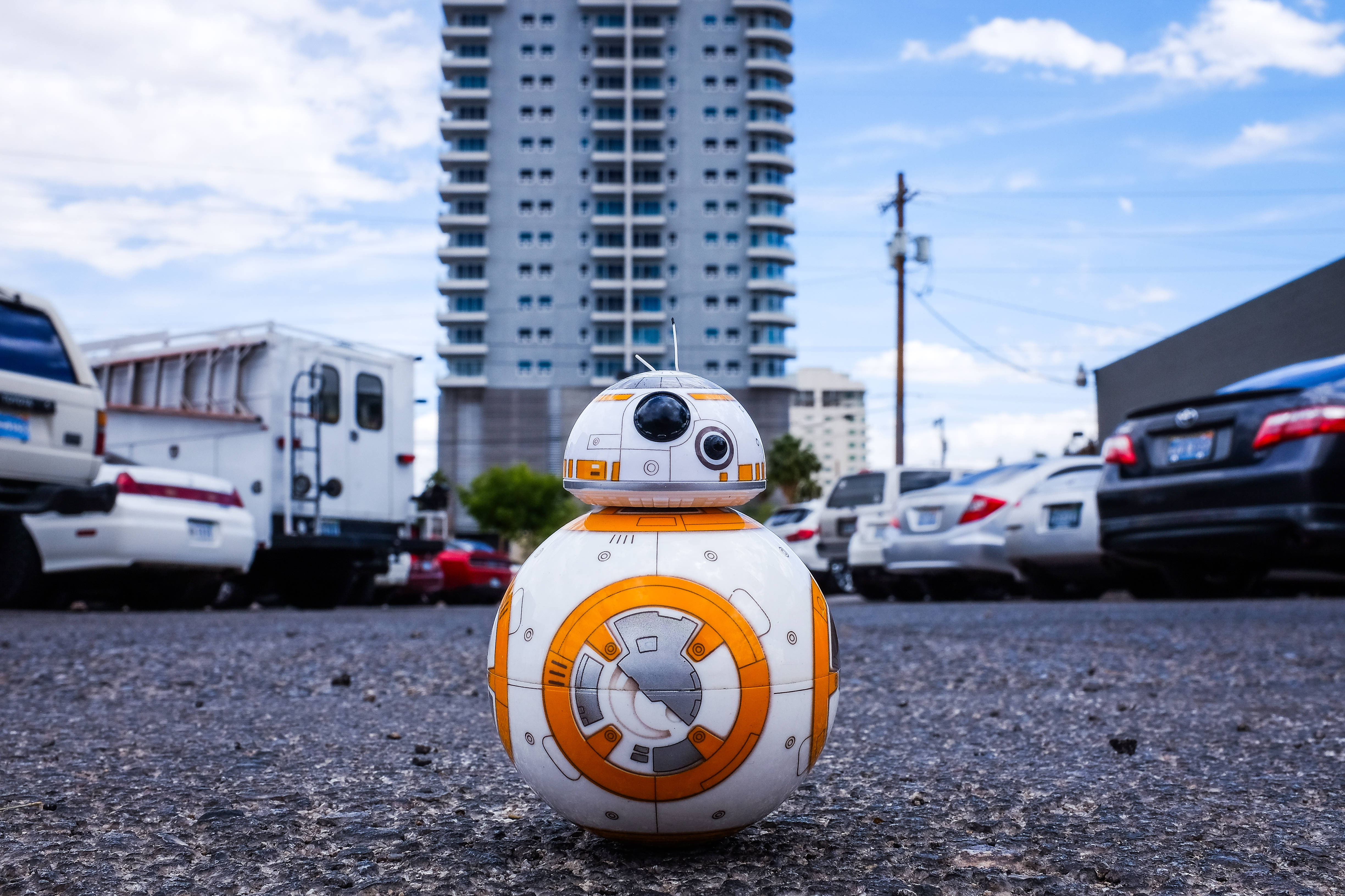 星際大戰 桌布 壁紙 — 一個BB-8機器人站在停車場的瀝青上，周圍有汽車和一棟高樓，天空部分多雲。