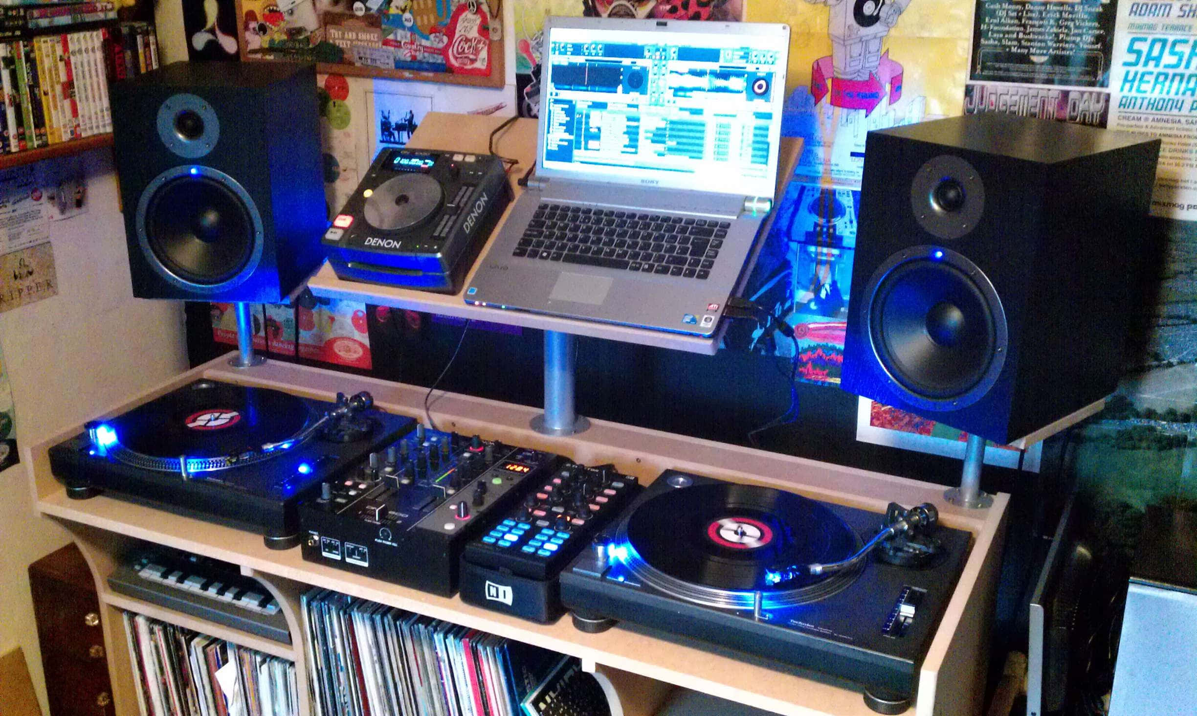 音樂 桌布 壁紙 — 一個DJ設置，有兩個唱盤、一個混音器、筆記型電腦和揚聲器，在雜亂的音樂室中發出藍光。