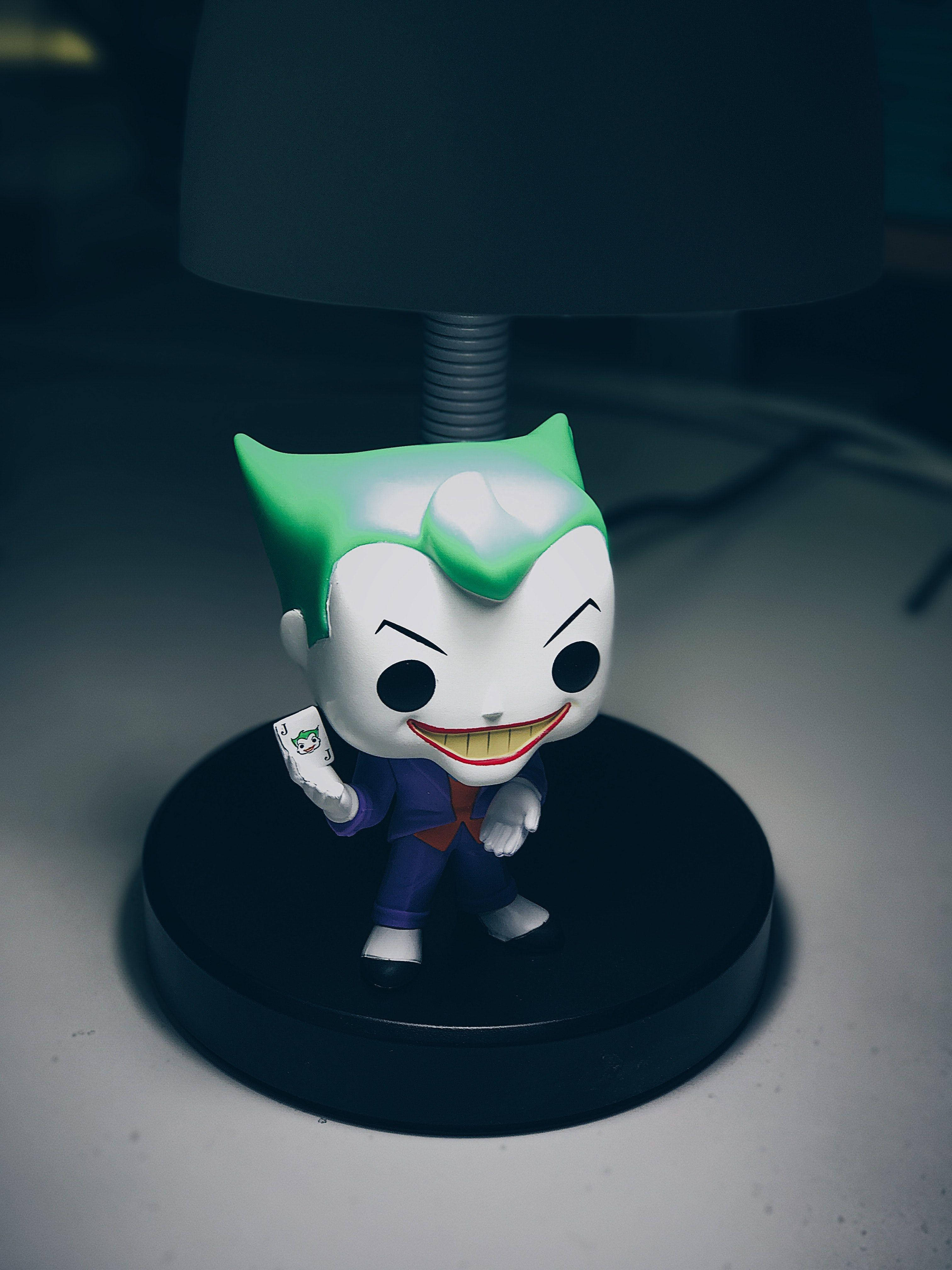 pop 桌布 壁紙 — 一個Funko Pop! 小丑公仔，綠色頭髮，紫色西裝，在白色表面上，暗燈下拿著一張小丑牌。