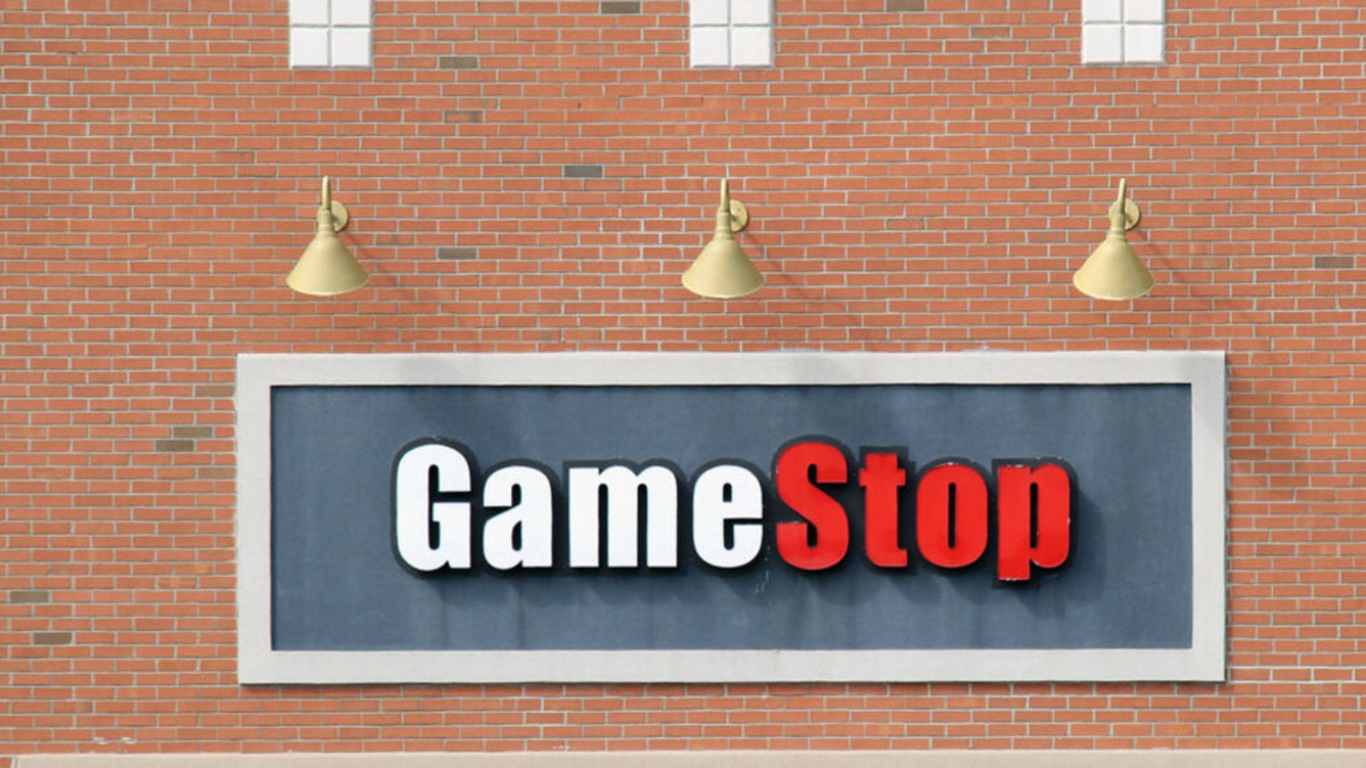 GameStop 桌布 壁紙 — 一個GameStop商店招牌，以粗體白色和紅色字體安裝在紅磚牆上，上方有三盞金色燈光。