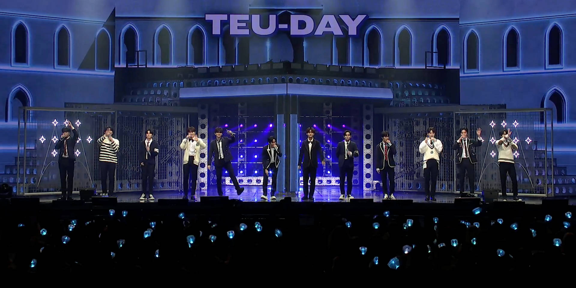 Treasure 桌布 壁紙 — 一個K-pop組合在一個宏偉的藍色燈光舞台上表演，帶有'TEU-DAY'標牌，粉絲們在下方舉著發光的藍色燈光。