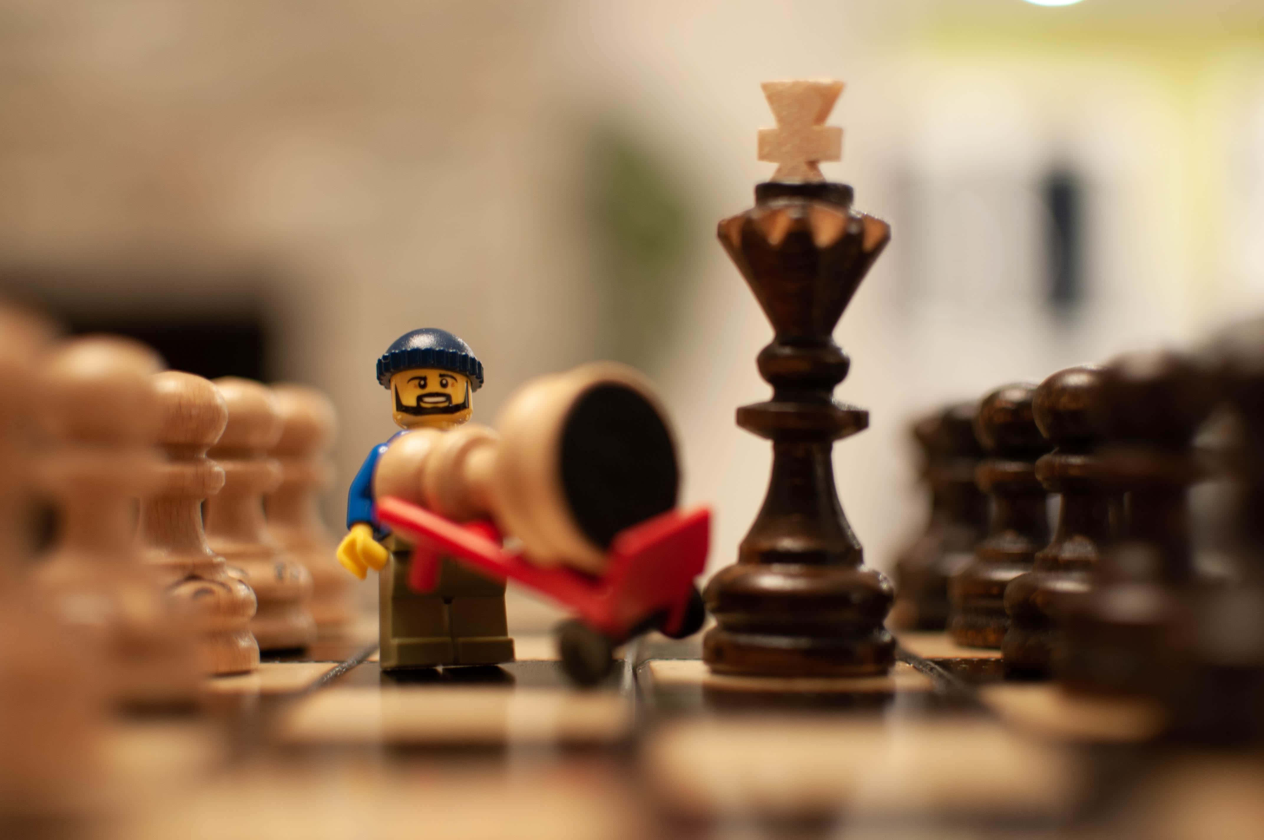 樂高 桌布 壁紙 — 一個LEGO迷你人偶在木製棋盤上推動一個倒下的棋子，前景中有一個巨大的國王棋子。
