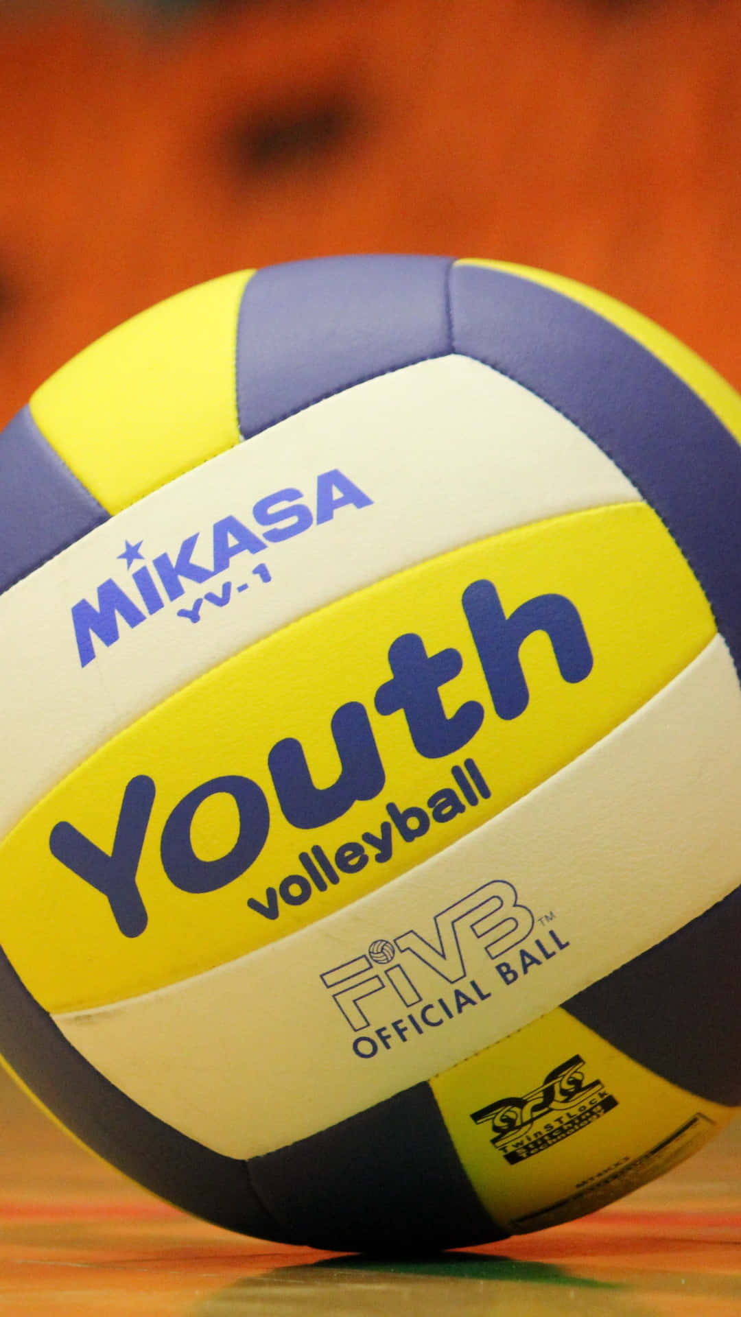 Volleyball Ball 桌布 壁紙 — 一個Mikasa Youth排球的特寫，有藍色、黃色和白色的面板，放在木製球場上。