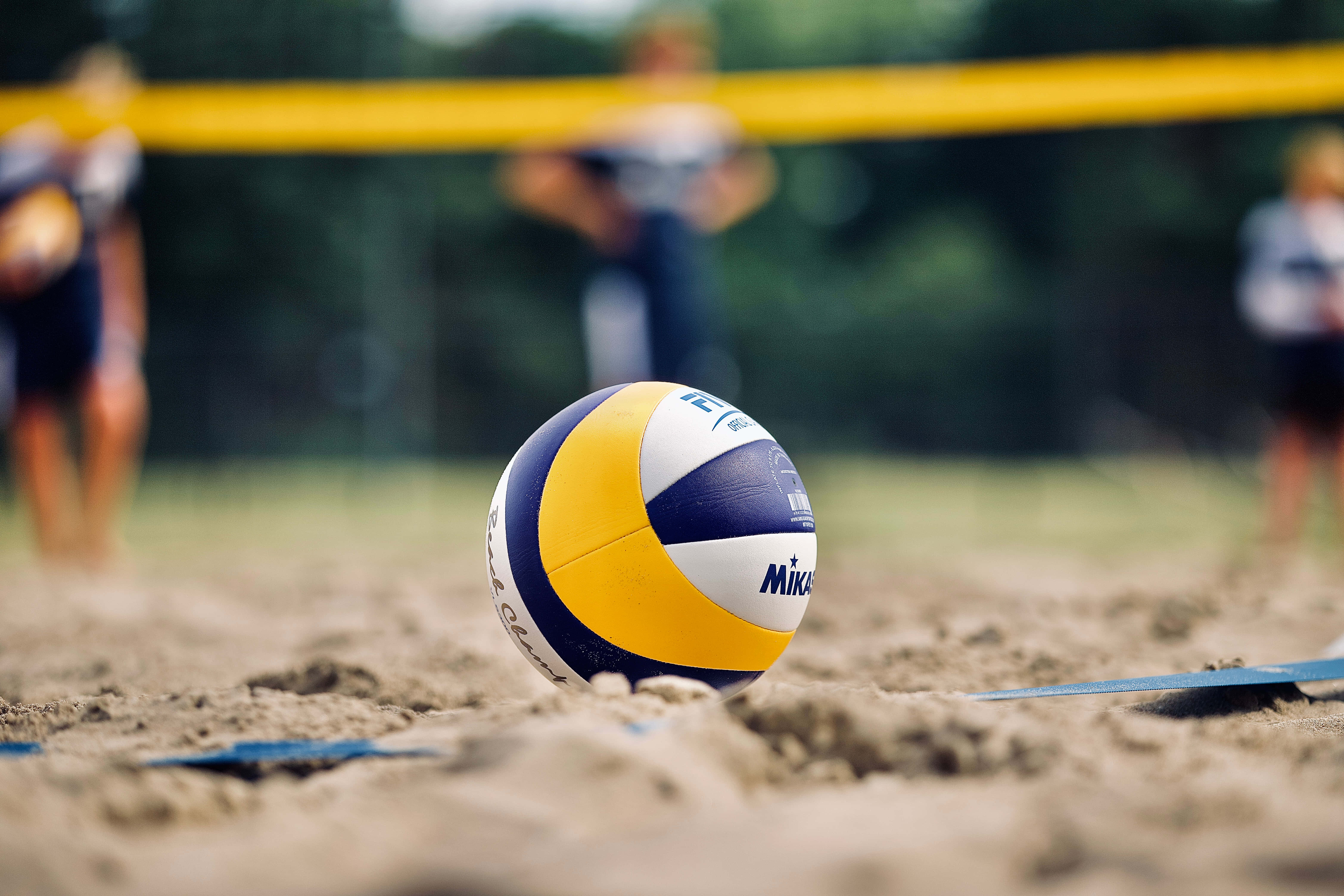 Volleyball Ball 桌布 壁紙 — 一個Mikasa排球擱在沙上，背景有模糊的沙灘排球選手和球網。