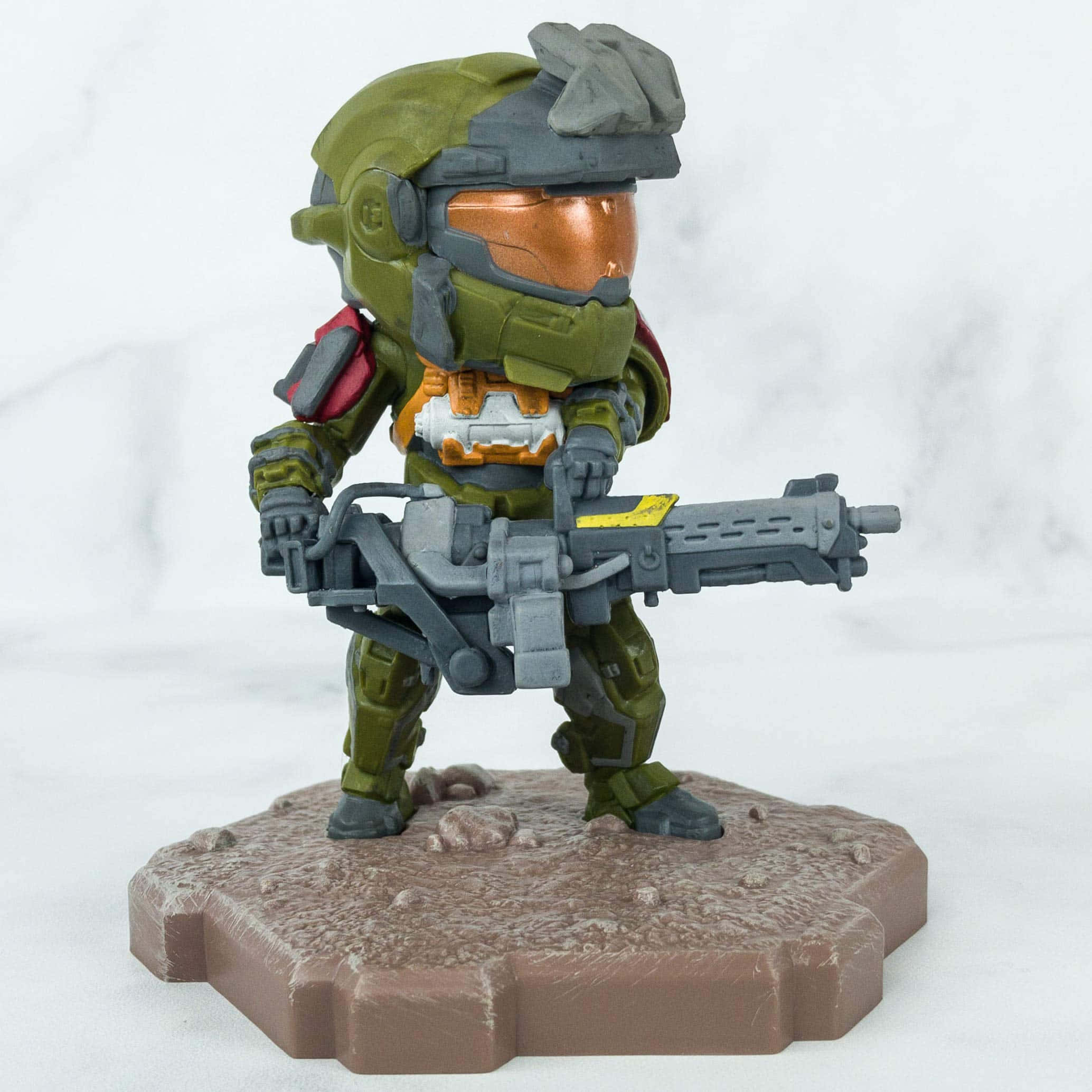 Jorge-052 桌布 壁紙 — 一個Q版Halo Master Chief公仔，身穿綠色盔甲，在白色大理石背景前的岩石底座上手持一把大槍。