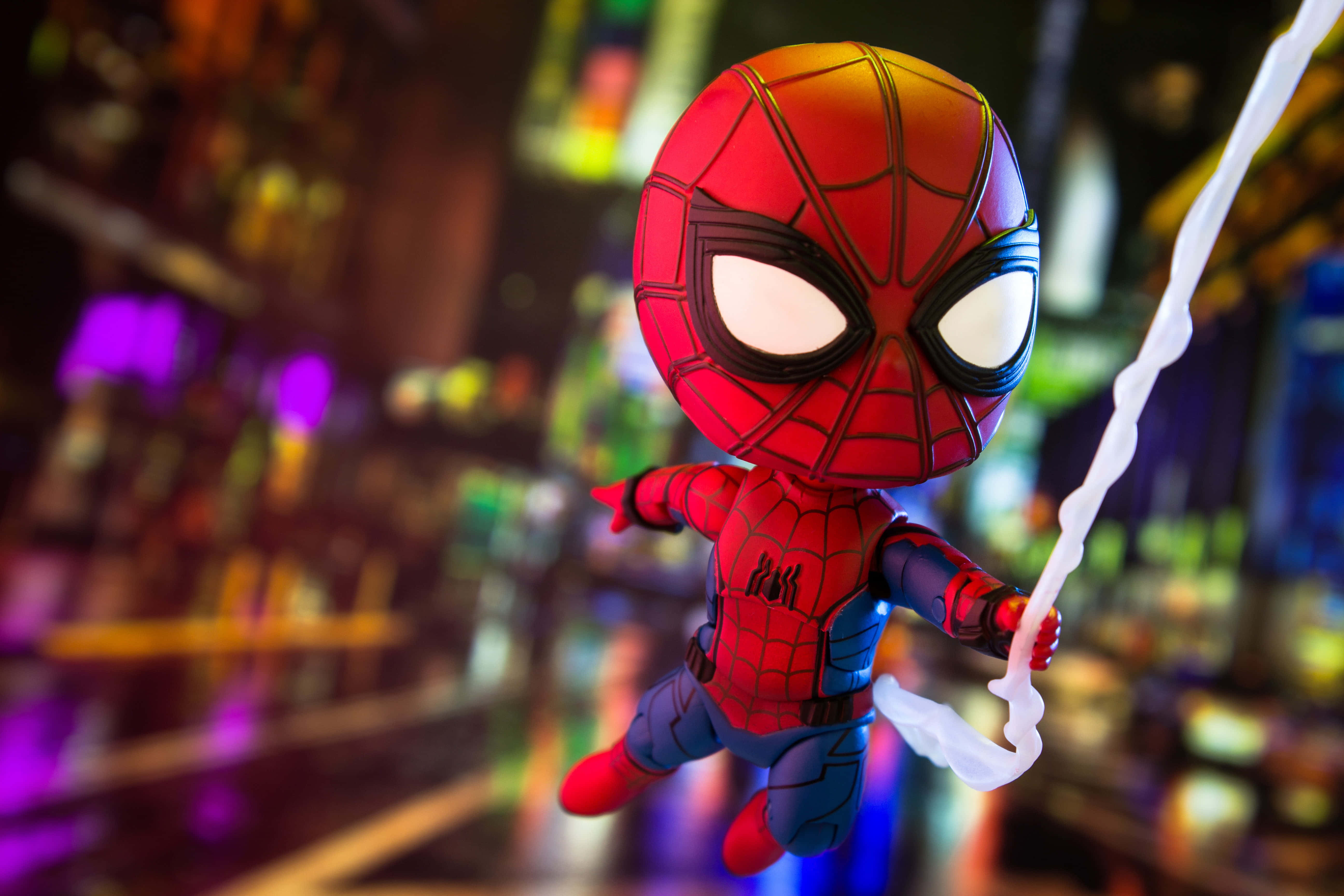 Spider-man Web Slinging 桌布 壁紙 — 一個Q版蜘蛛俠人偶在夜晚的霓虹燈城市景觀中擺盪，帶有發光的蜘蛛網線。