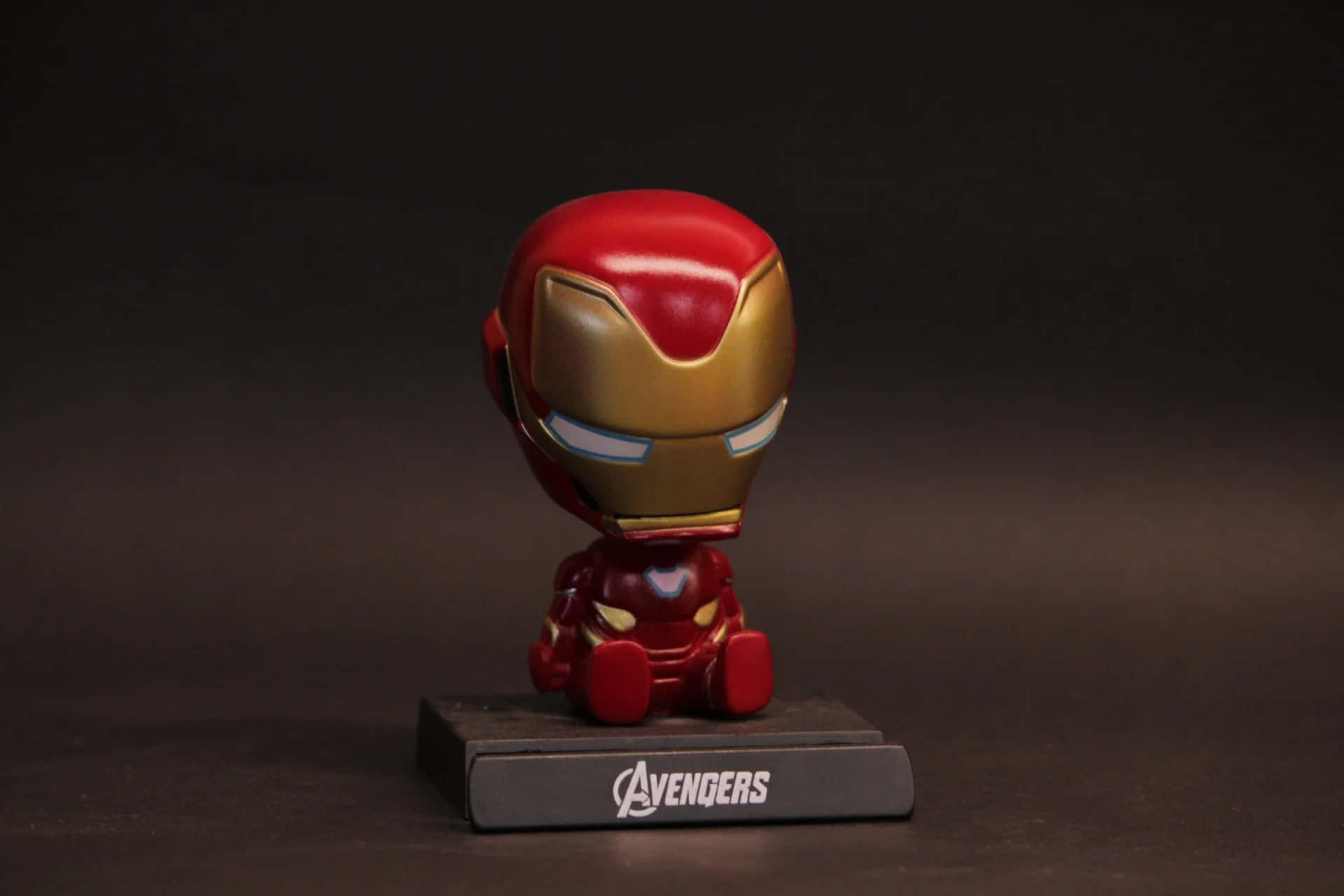 Iron Man Bobbleheads 桌布 壁紙 — 一個Q版風格的鋼鐵人人偶坐在黑色復仇者聯盟底座上，背景為深色攝影棚。