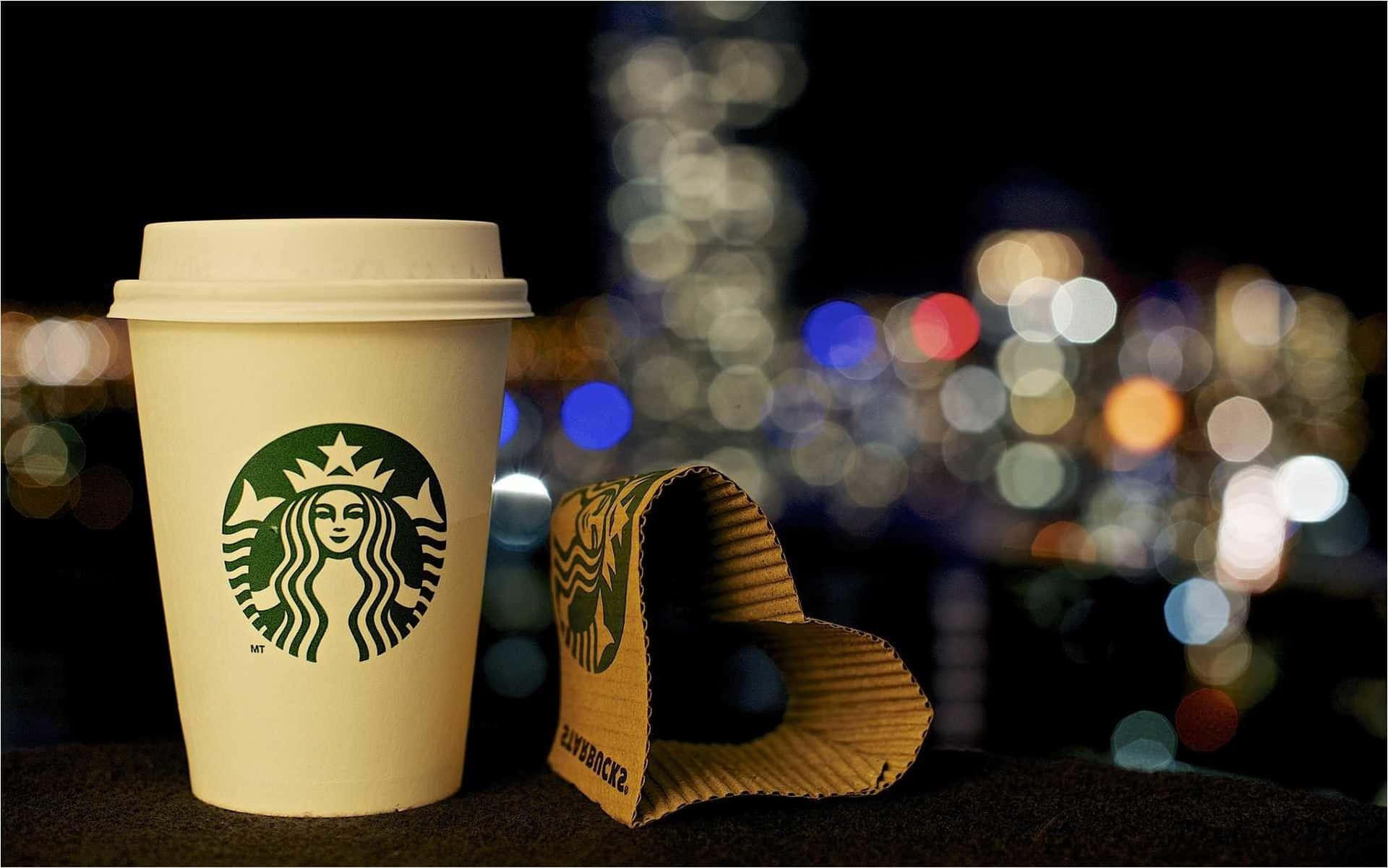 星巴克 桌布 壁紙 — 一個Starbucks咖啡杯及其杯套放在窗台上，背景是充滿活力的散景城市夜景天際線。