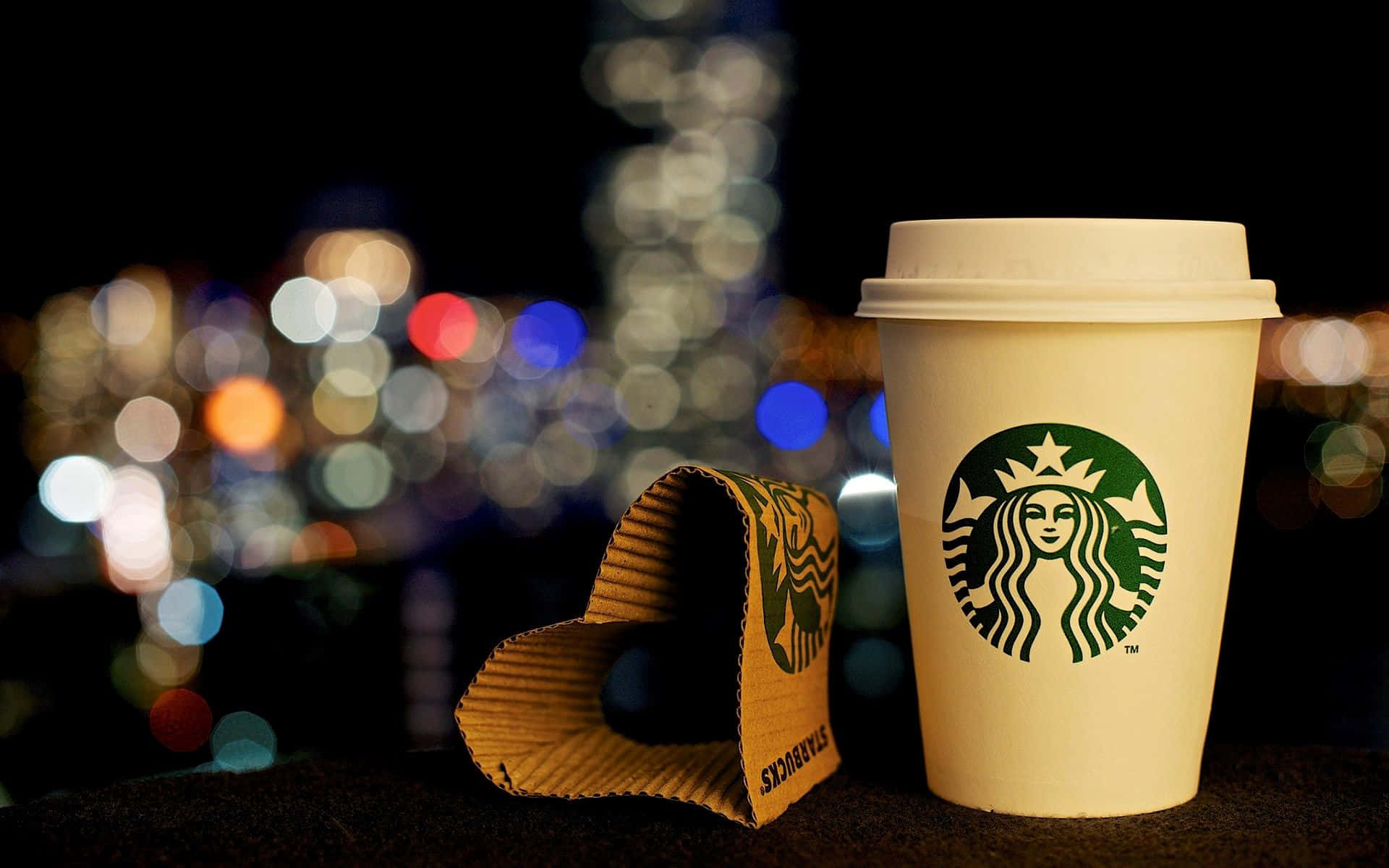 星巴克 桌布 壁紙 — 一個Starbucks咖啡杯和杯套放在窗台上，背景是夜晚充滿活力的散景城市景觀。