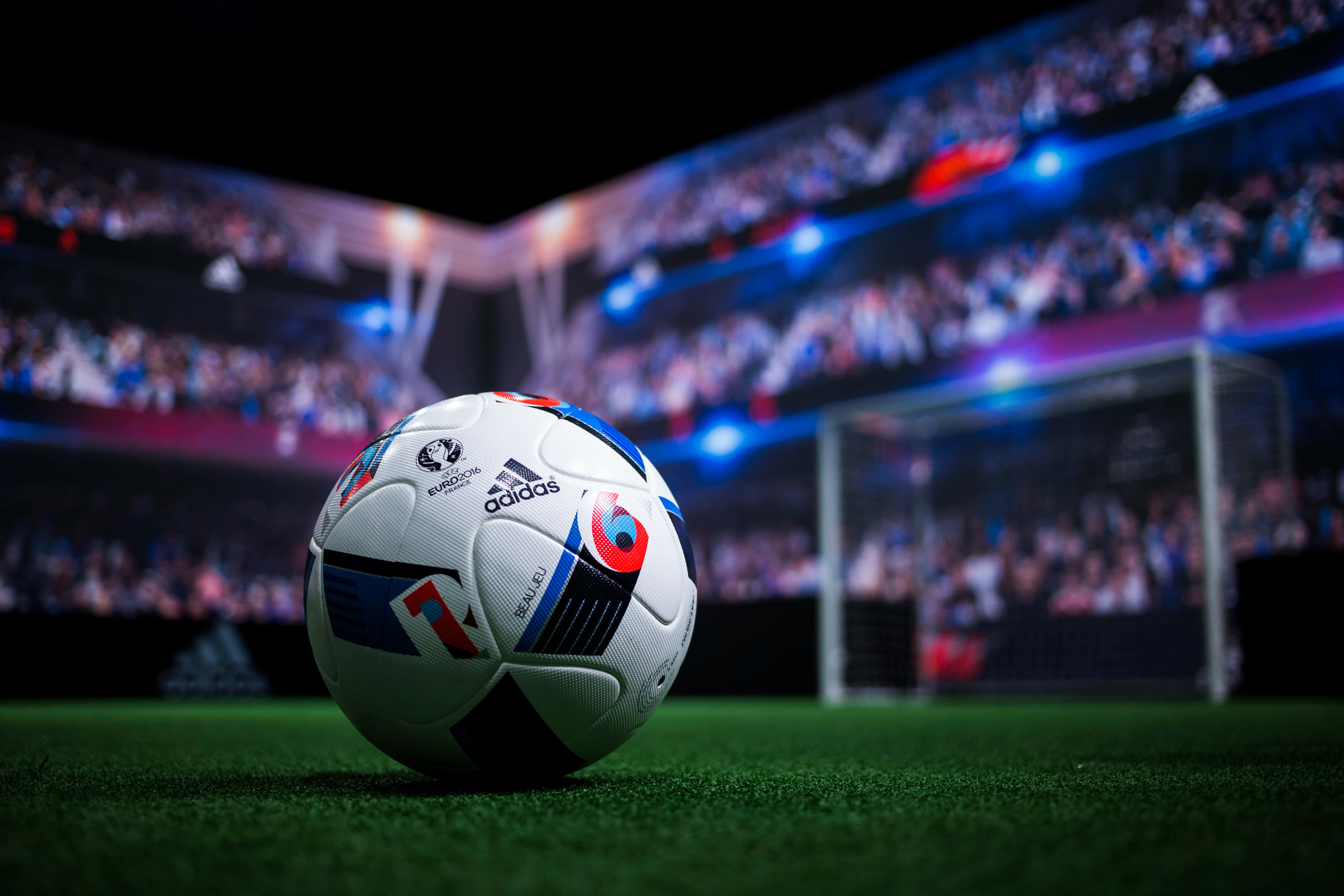 足球 桌布 壁紙 — 一個UEFA Euro 2020足球放在充滿活力的綠色草地上，模糊的體育場人群和球門網在戲劇性的藍色燈光下閃耀。