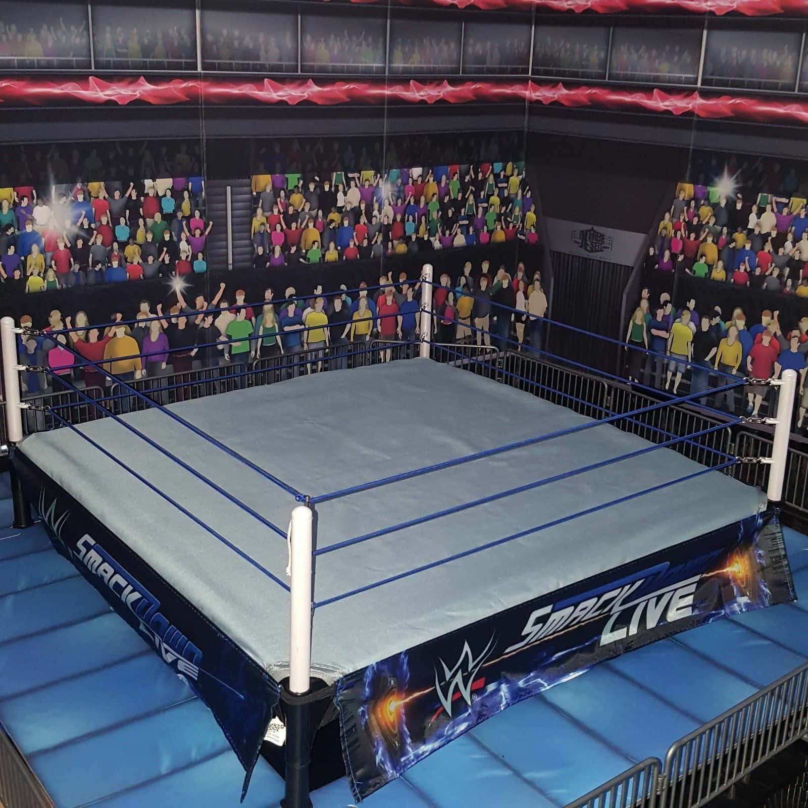 Wrestling Ring 桌布 壁紙 — 一個WWE SmackLive摔角擂台，配有藍色繩索和墊子，周圍是昏暗場館中歡呼的人群壁畫。