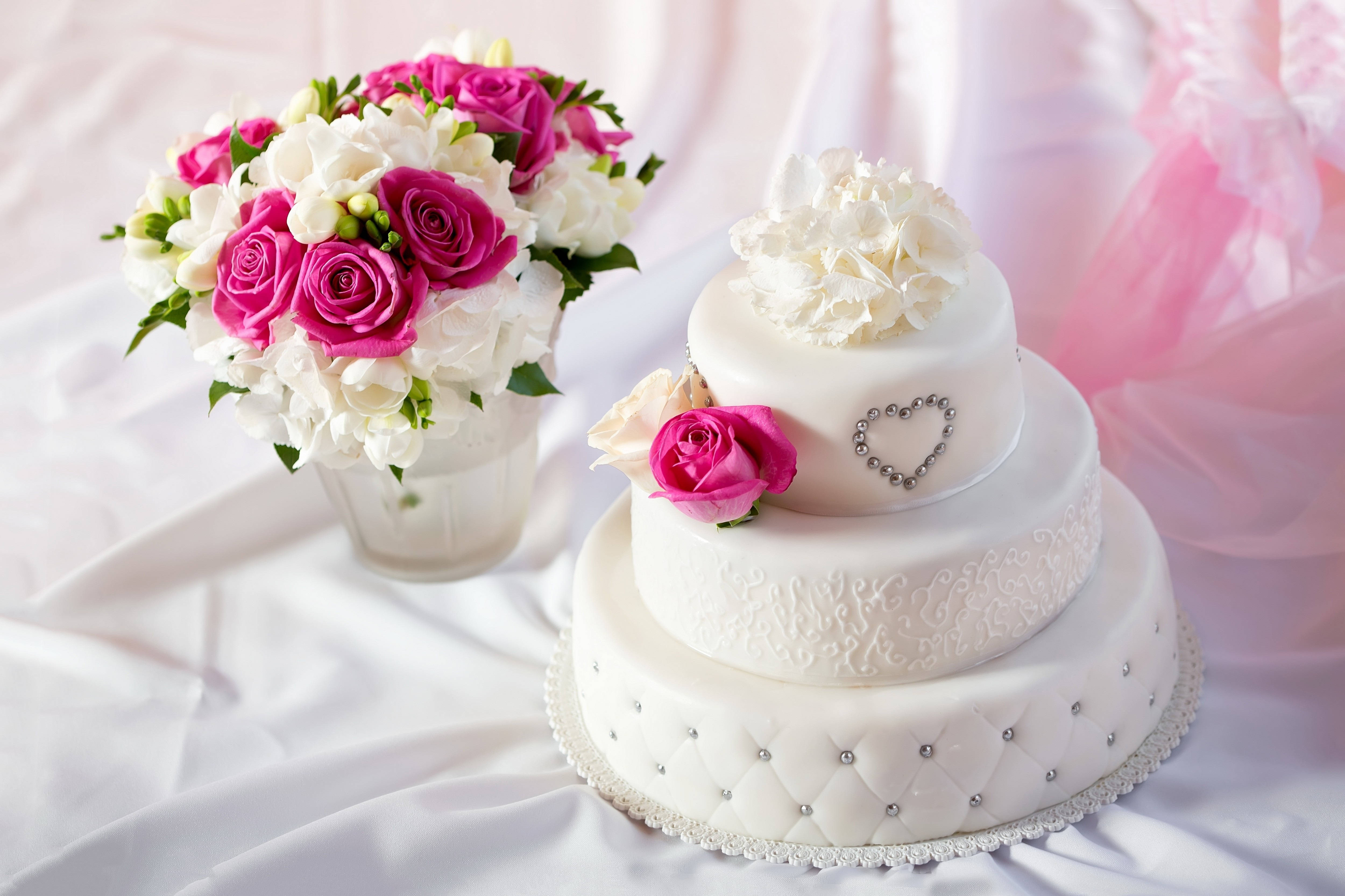 Wedding 桌布 壁紙 — 一個三層白色結婚蛋糕，裝飾著粉紅玫瑰和銀色珠子，旁邊放著一束相配的花束，放在白色垂褶布上。