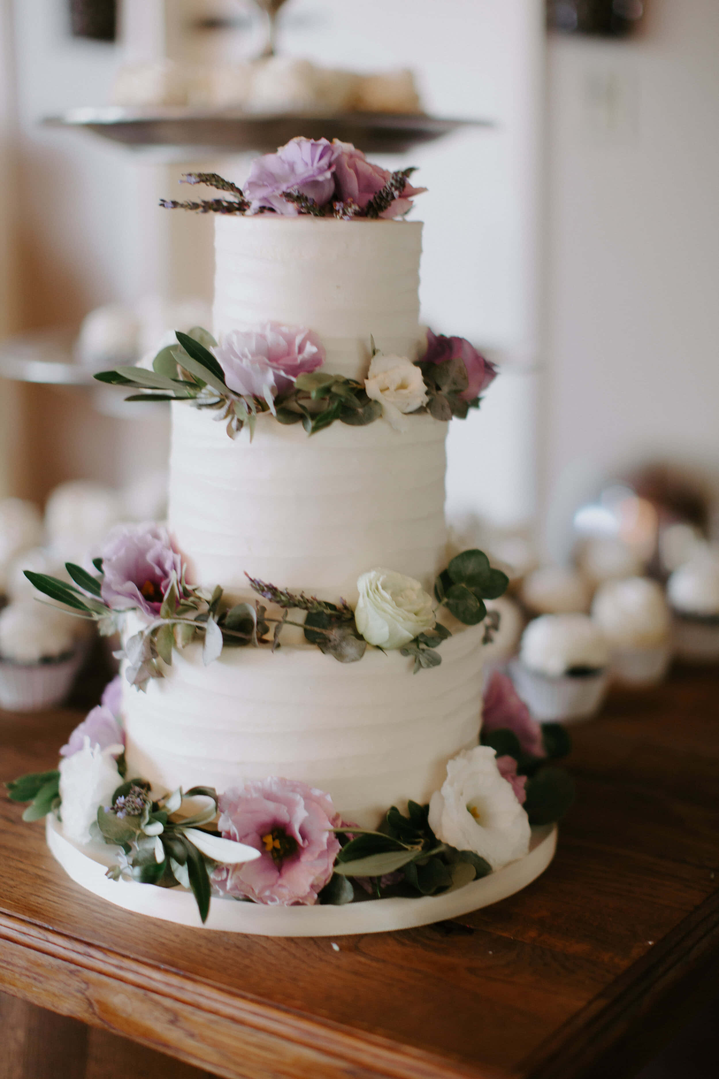 Cake Fondant 桌布 壁紙 — 一個三層白色結婚蛋糕，飾以柔和的粉紅色和白色花朵及綠葉，優雅地放在木桌上。