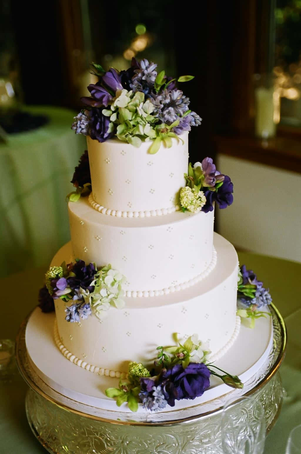 Wedding Cake 桌布 壁紙 — 一個三層白色結婚蛋糕，飾以紫色和綠色花朵，優雅地放置在一個水晶架上。
