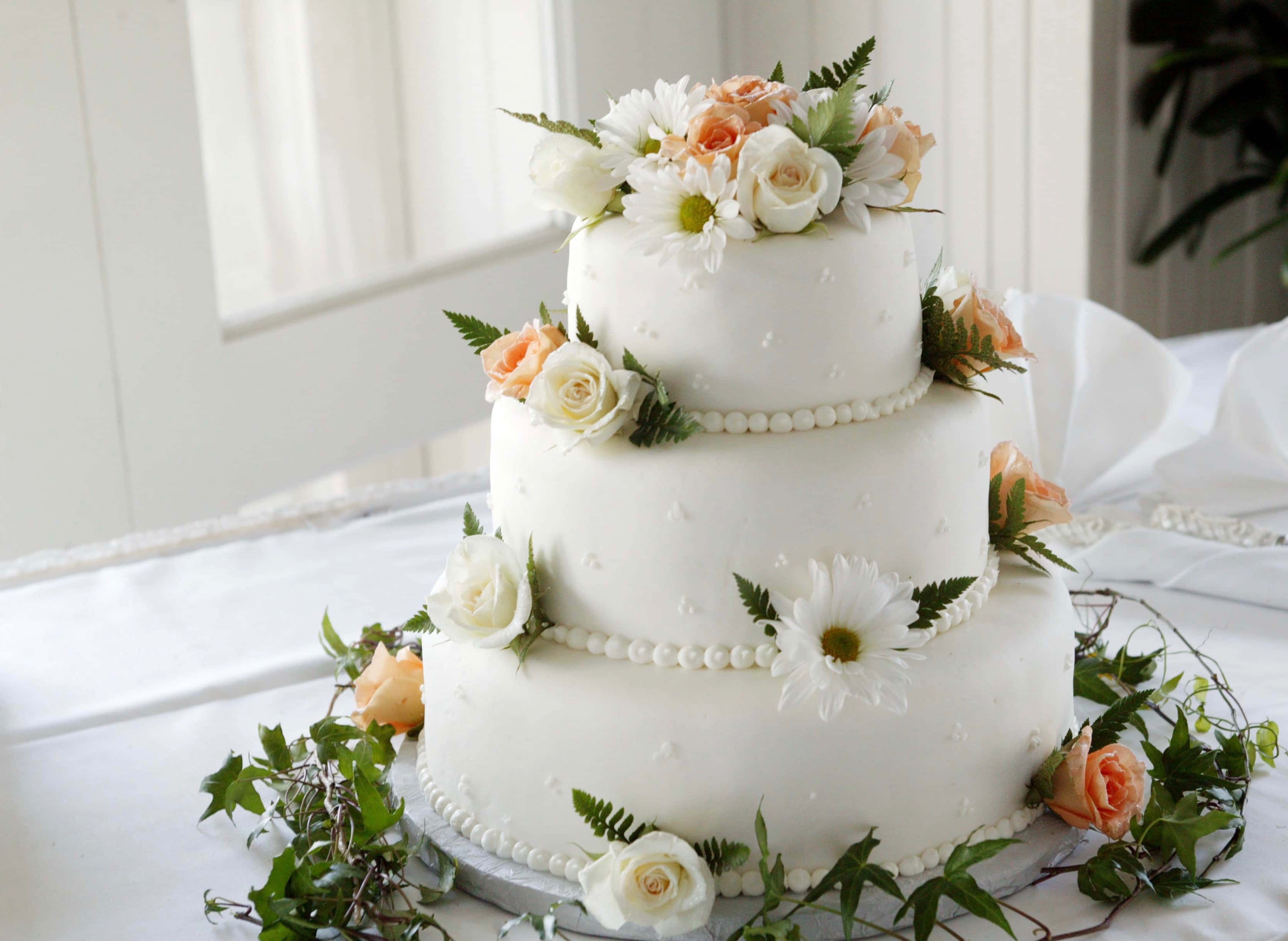 Floral Cake 桌布 壁紙 — 一個三層白色結婚蛋糕，飾有蜜桃色和白色玫瑰及綠葉，優雅地放在白色桌布上。
