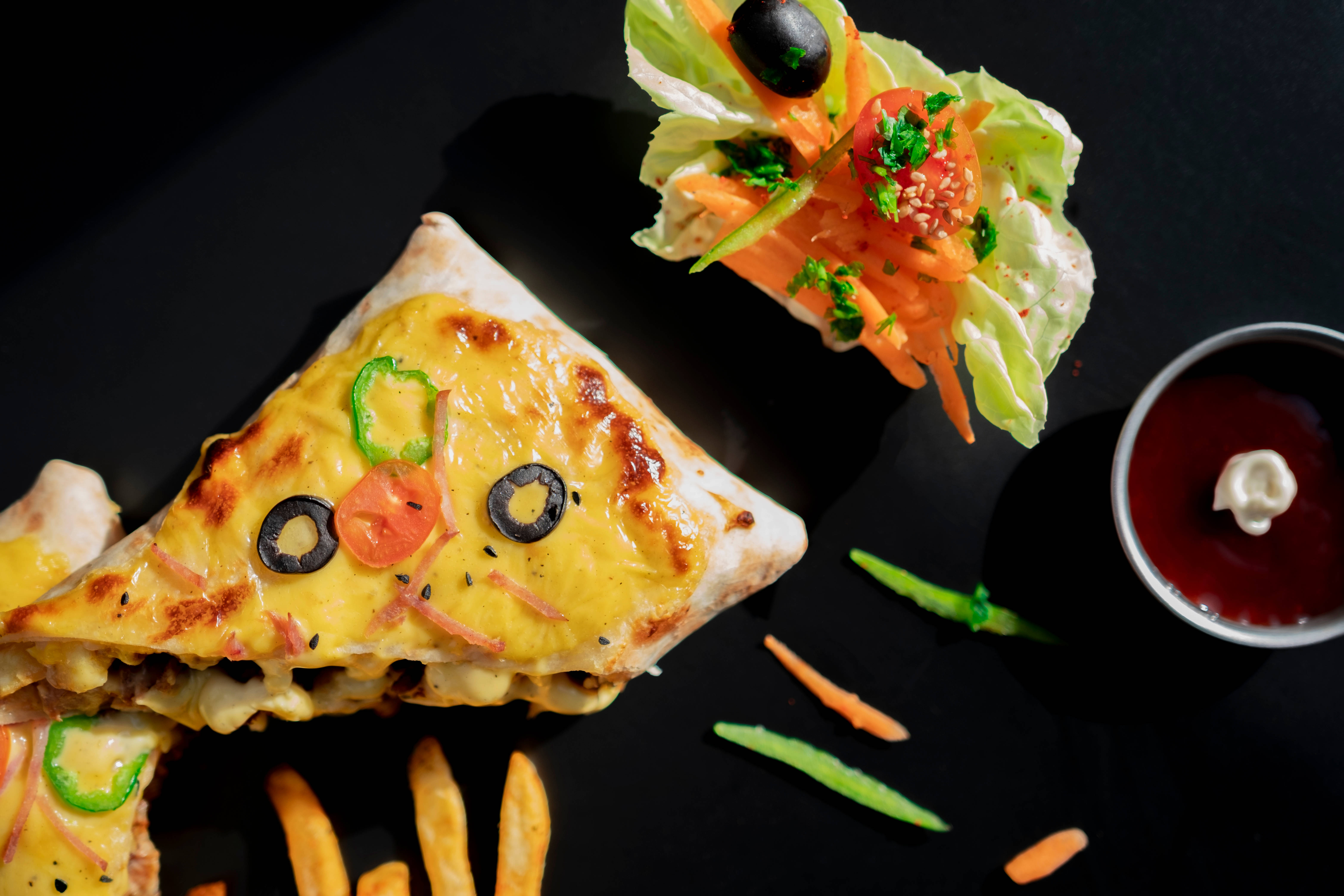 4K Food 桌布 壁紙 — 一個三角形起司漢堡披薩，上面用橄欖和墨西哥辣椒排列成一張臉，旁邊放著新鮮沙拉和沾醬，放在黑色盤子上。