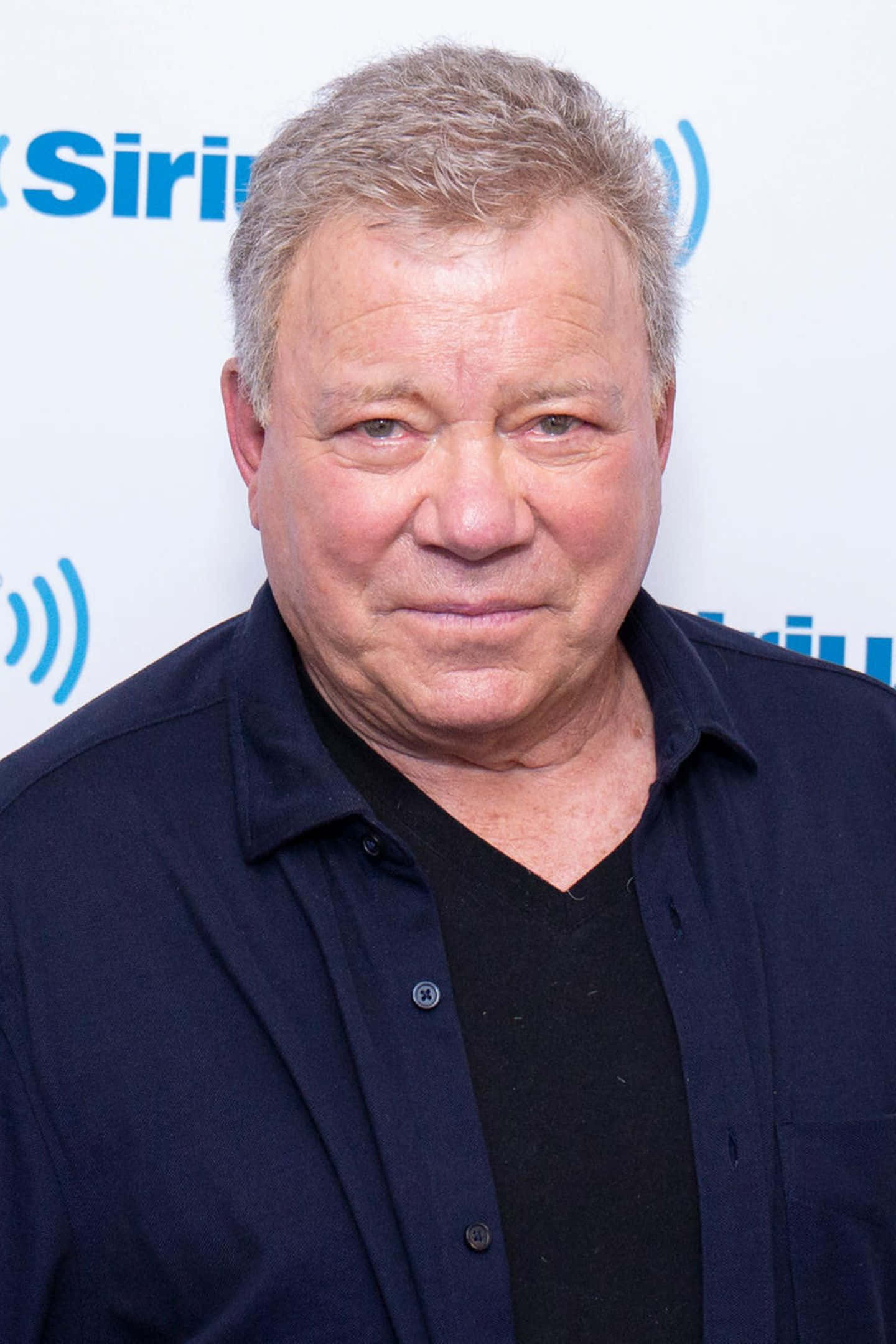 william shatner 桌布 壁紙 — 一個中年男子的特寫肖像，他有灰白頭髮，在SiriusXM活動中穿著一件深藍色鈕扣襯衫，內搭黑色V領。