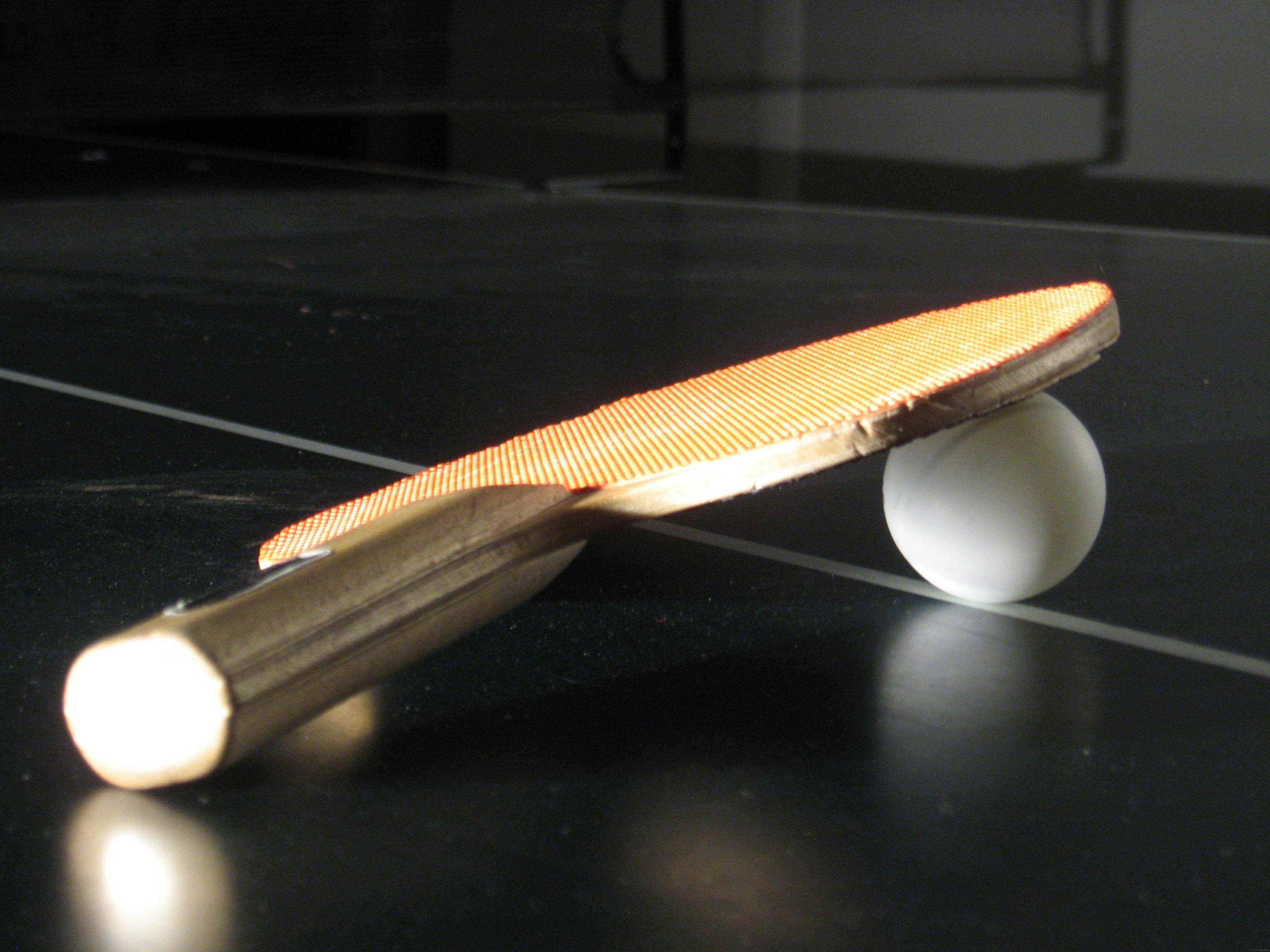 Table Tennis 桌布 壁紙 — 一個乒乓球拍和一個乒乓球放在深色桌子上，溫暖的方向性燈光投射出柔和的陰影。
