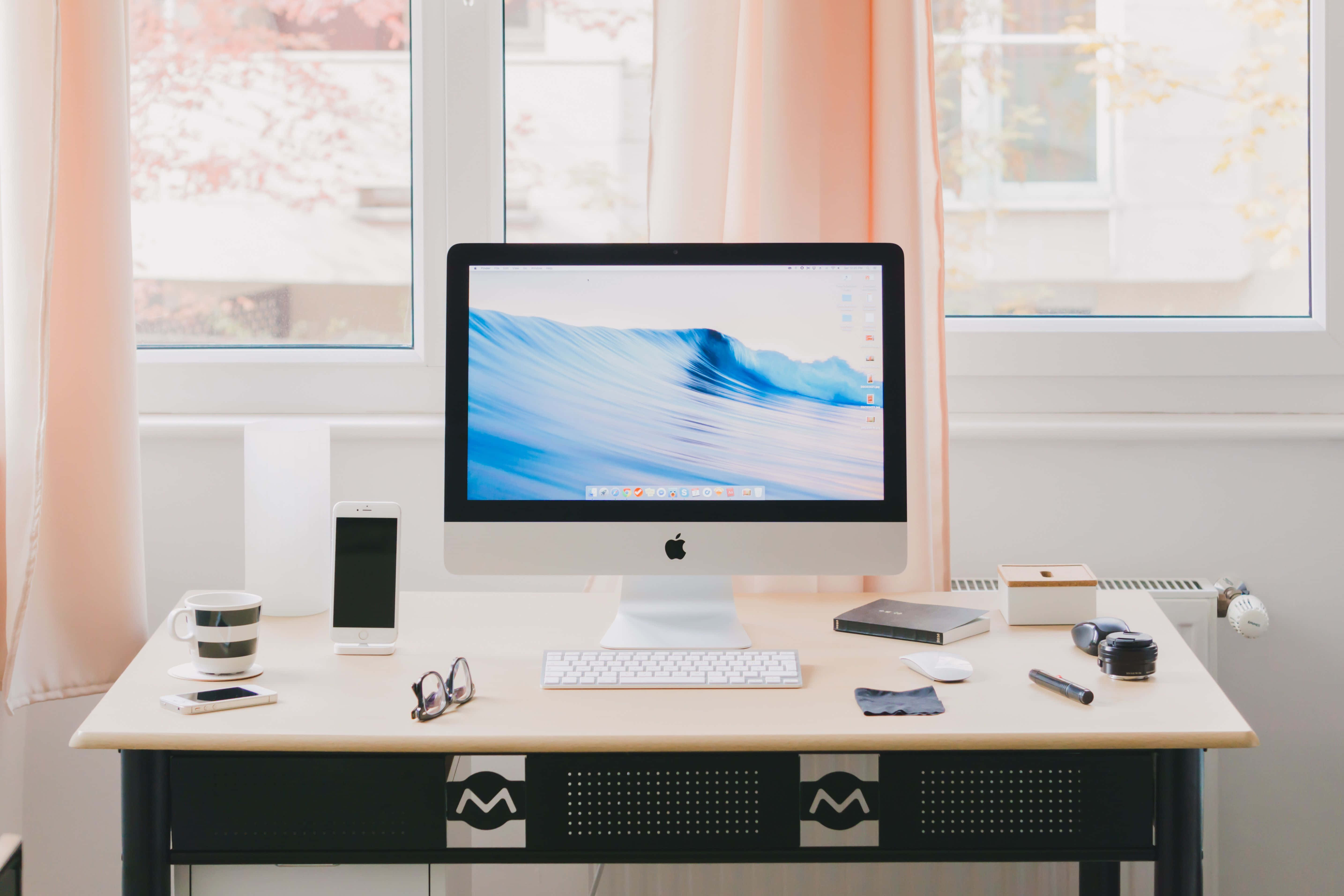 Working From Home 桌布 壁紙 — 一個乾淨的現代辦公桌設置，配備一部顯示藍色波浪桌面的iMac，周圍有科技小工具和一個杯子，在柔和窗光下。