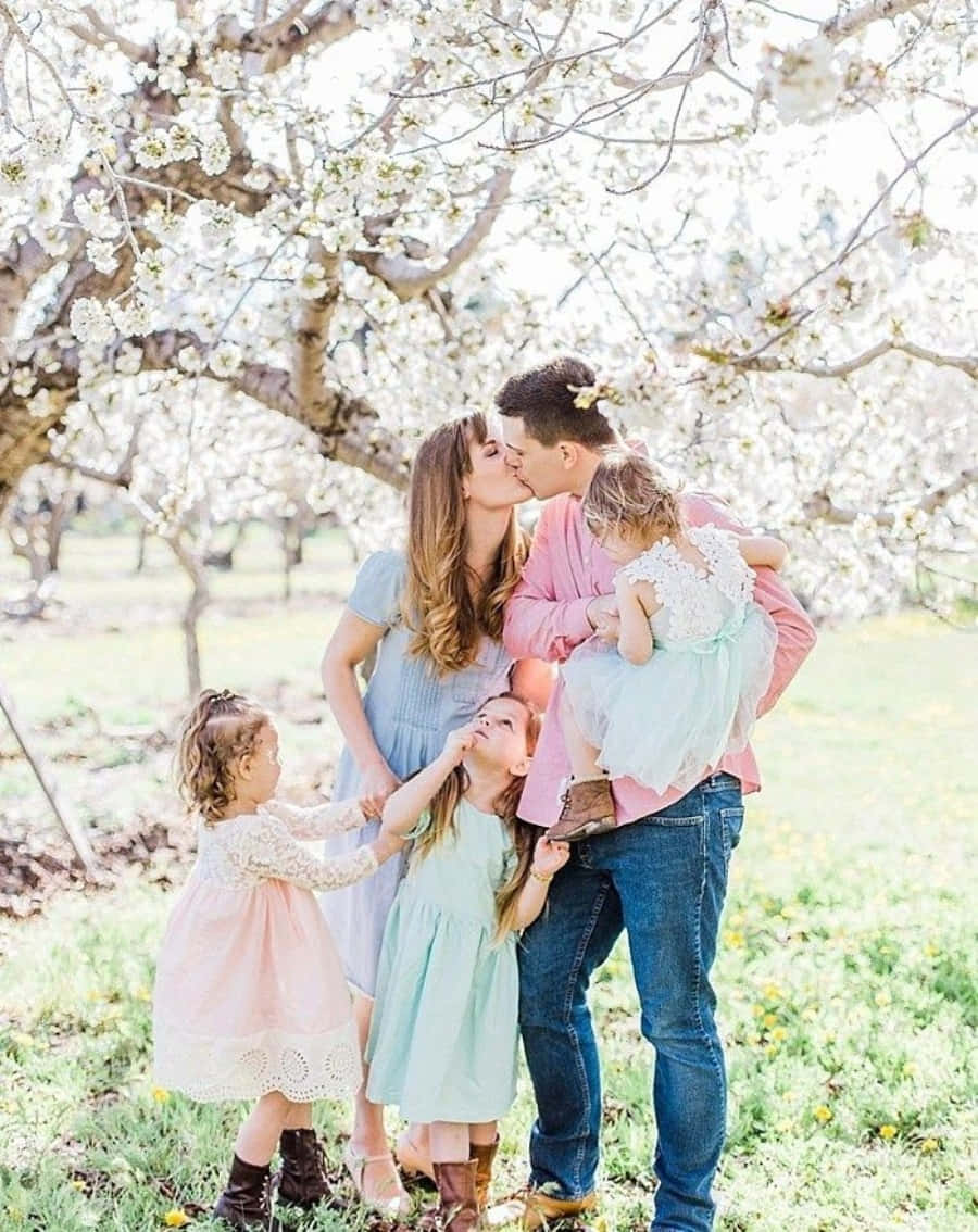 Spring Family 桌布 壁紙 — 一個五口之家在陽光明媚的日子裡，在盛開的白櫻花樹冠下分享一個溫柔的吻。