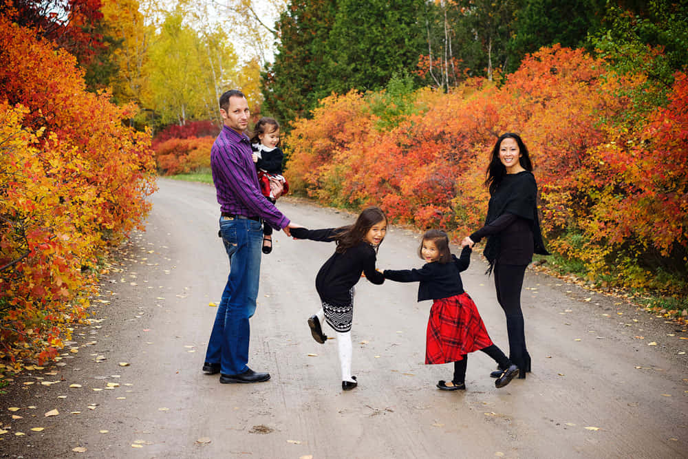 Fall Family 桌布 壁紙 — 一個五口之家快樂地手牽手，沿著一條蜿蜒的秋日小徑跳舞，小徑兩旁是火紅的橙色和紅色樹葉。