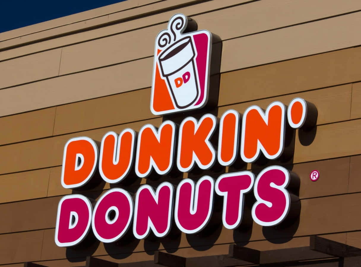 Dunkin Donuts 桌布 壁紙 — 一個亮橙色和粉紅色的Dunkin' Donuts招牌，帶有咖啡杯標誌，安裝在米色建築物外牆上，背景是晴朗的藍天。