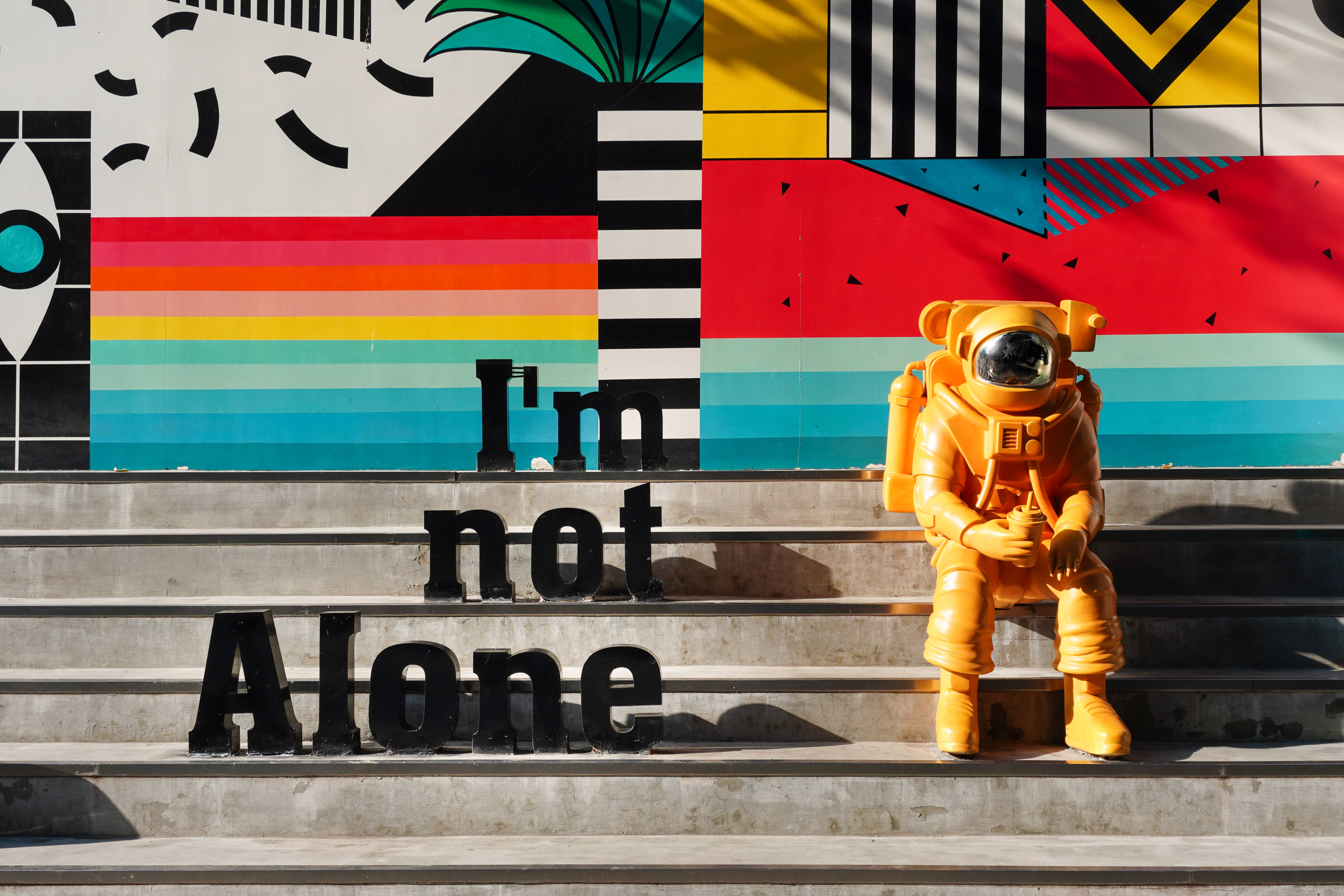Forever Alone 桌布 壁紙 — 一個亮橙色太空人雕像坐在混凝土樓梯上，前面是一幅色彩繽紛的幾何壁畫，上面有粗體黑色字寫著「I'm not Alone」。