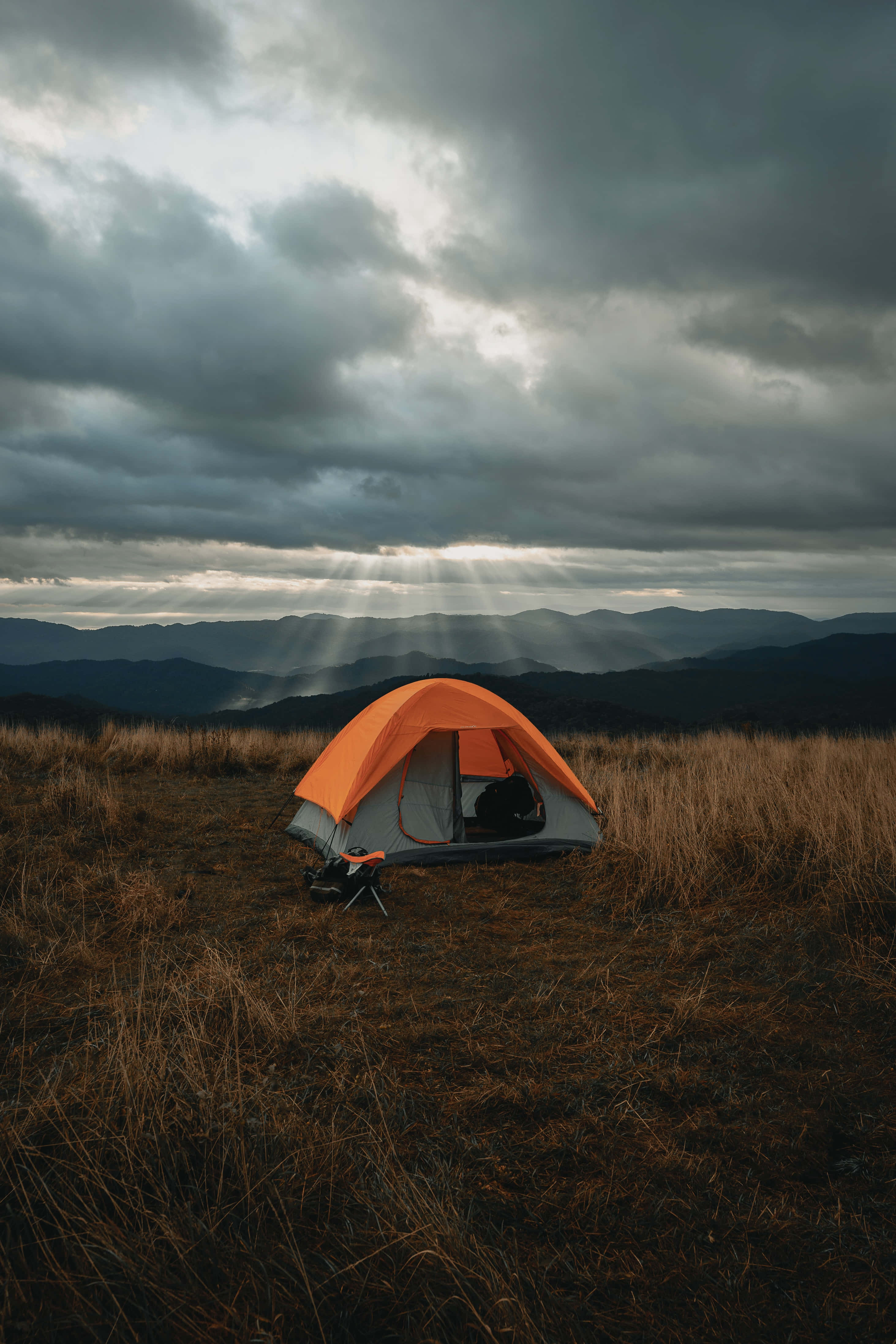 Camping 桌布 壁紙 — 一個亮橙色的帳篷獨自矗立在長滿草的山頂上，戲劇性的陽光穿透遠處山脈上方黑暗的暴風雲。