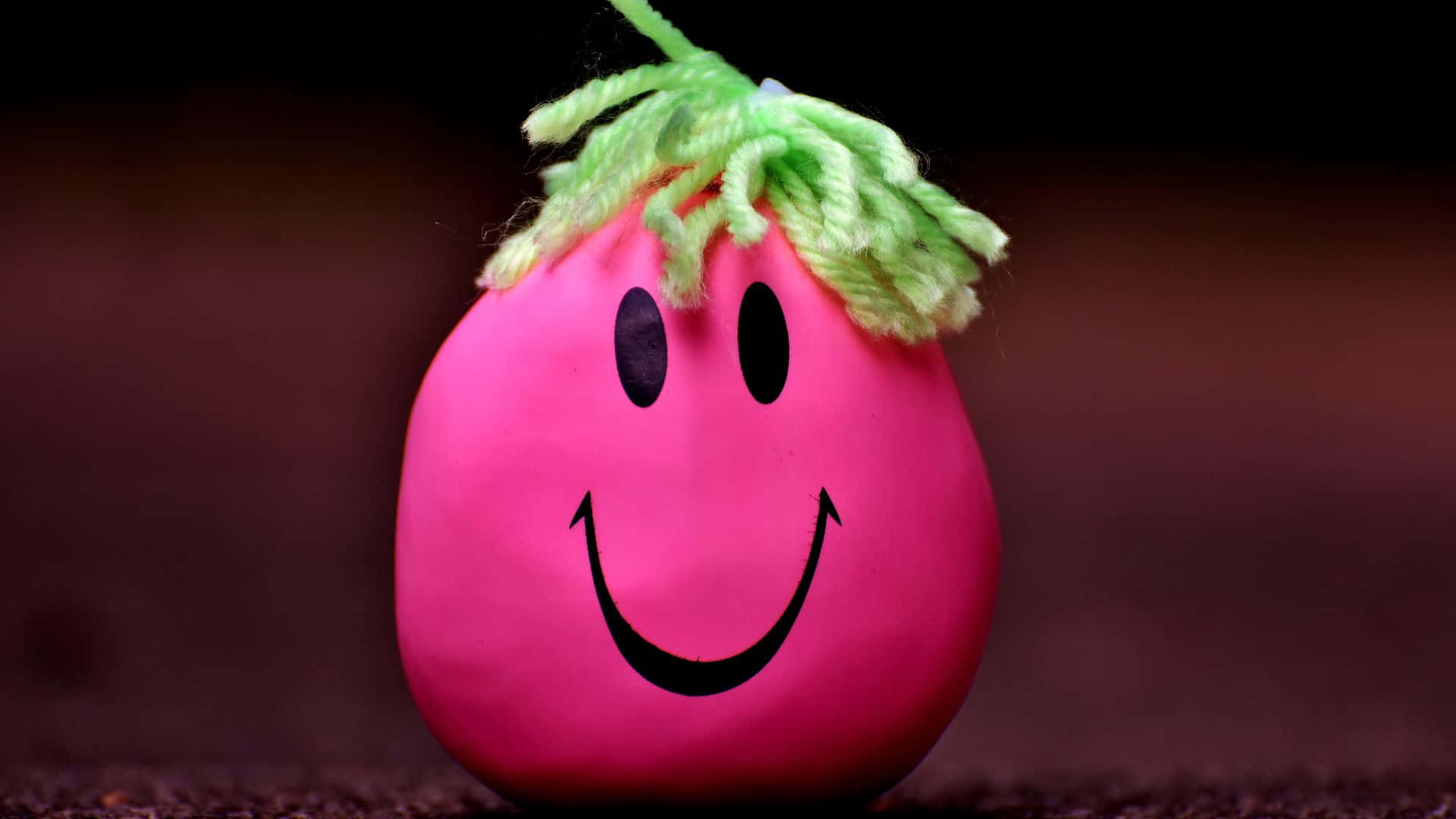 Smiley 桌布 壁紙 — 一個亮粉紅色蛋形玩具，帶有綠色毛線假髮和簡單的黑色笑臉，放在模糊的深色背景前。