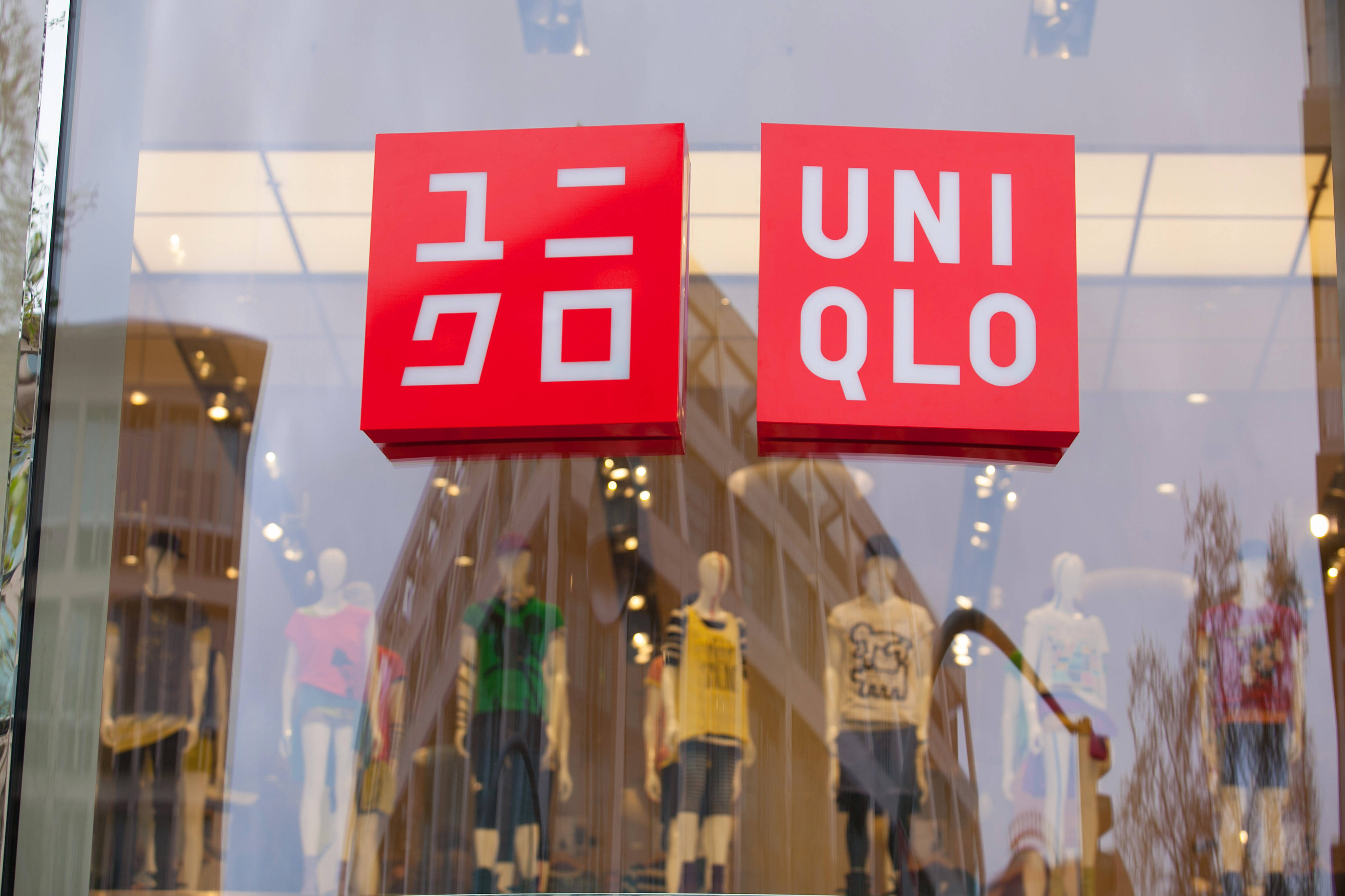 Uniqlo 桌布 壁紙 — 一個亮紅色的Uniqlo標誌懸掛在玻璃櫥窗內穿著色彩繽紛休閒服飾的人體模型上方。