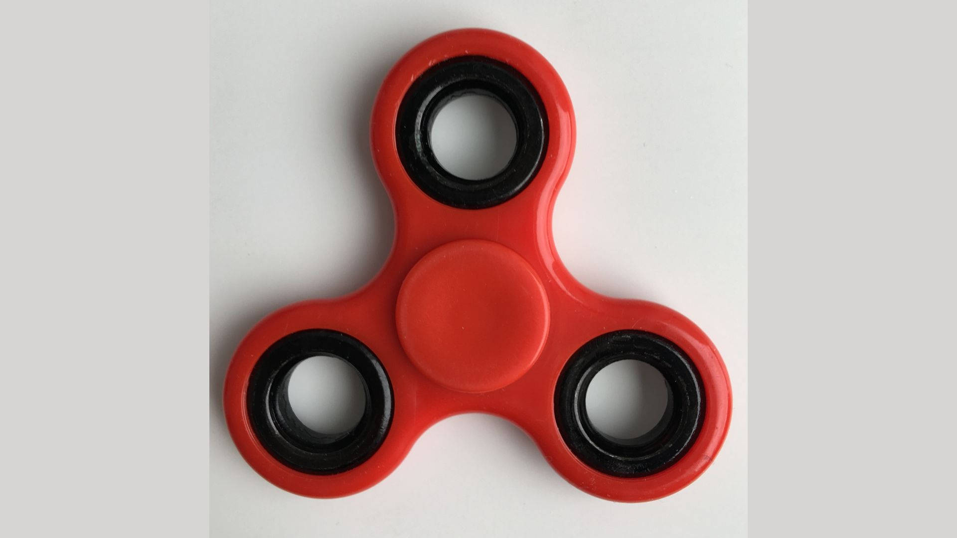 toys 桌布 壁紙 — 一個亮紅色的三叉指尖陀螺，帶有黑色軸承，放置在純白色表面上。