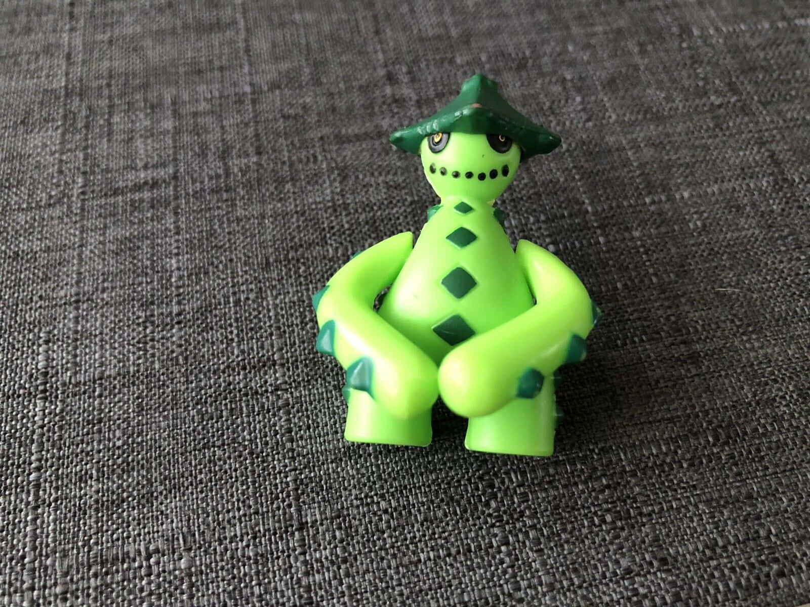 3D 桌布 壁紙 — 一個亮綠色塑料人偶戴著圓錐形帽子，盤腿坐在灰色紋理布料表面上。