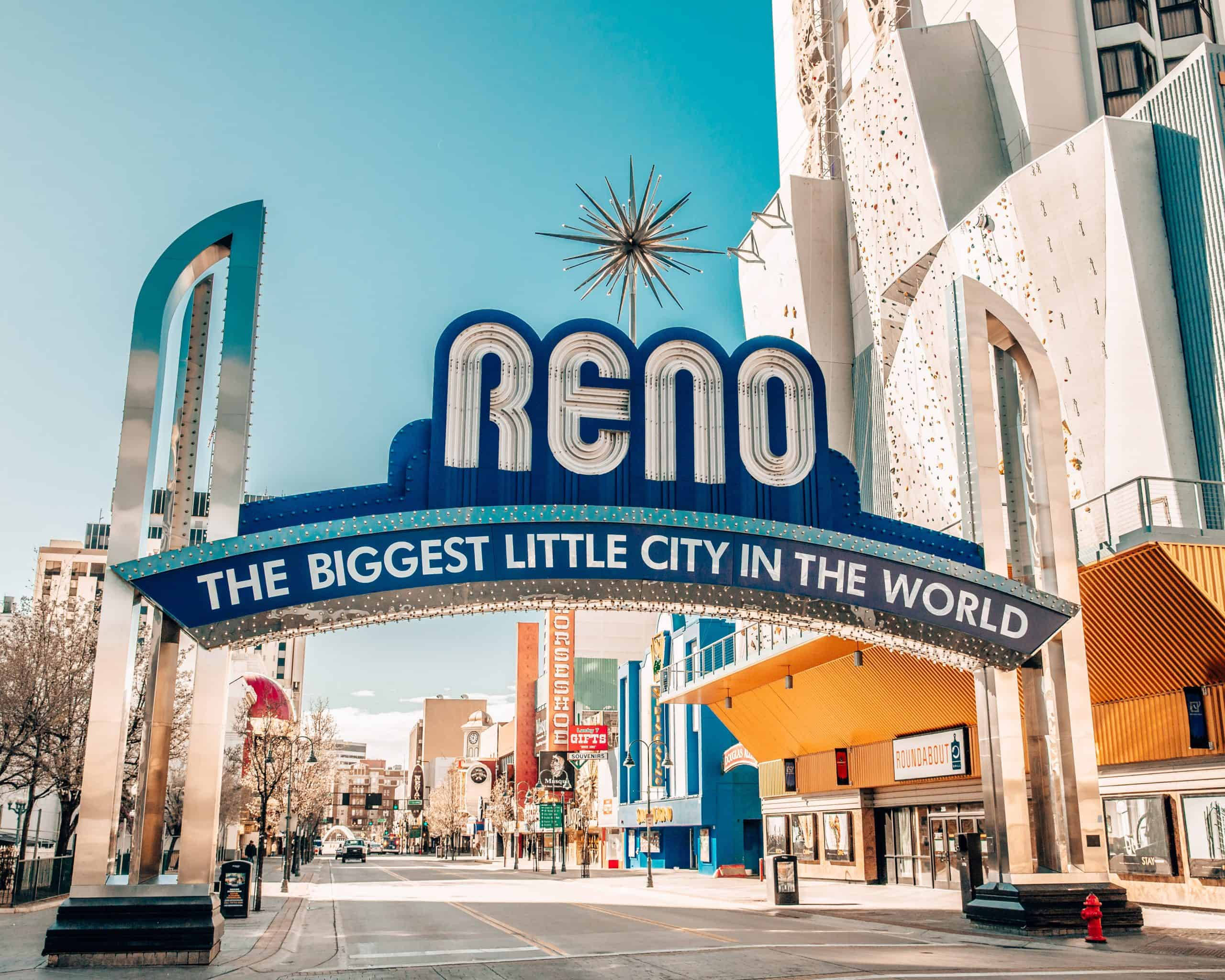 Arch 桌布 壁紙 — 一個亮藍色的「RENO」標誌拱形橫跨在陽光明媚的城市街道上，街道上有現代建築和一個星爆雕塑，背景是晴朗的天空。