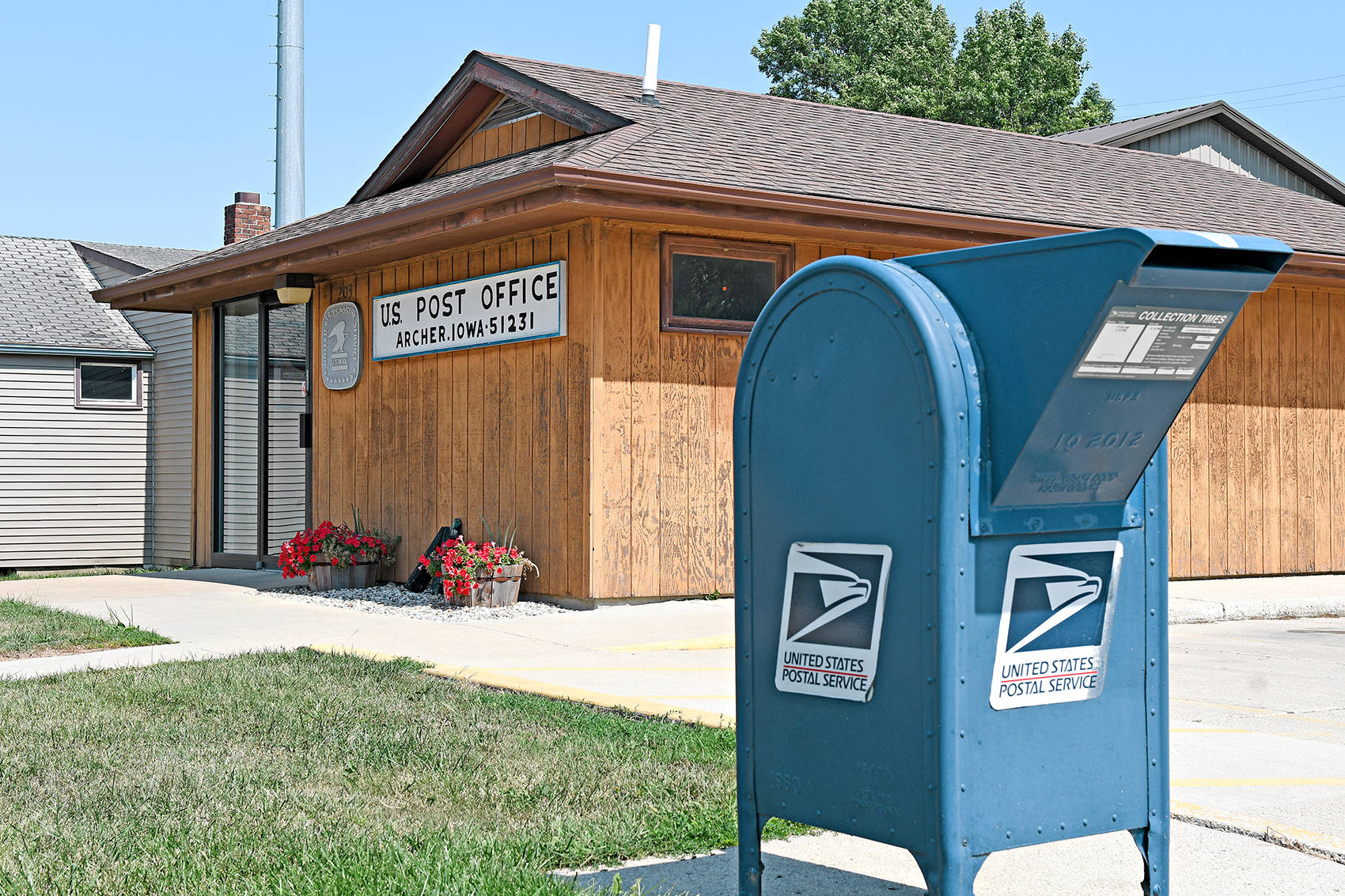 Post Office 桌布 壁紙 — 一個亮藍色的USPS郵箱在陽光明媚的日子裡，站在木質郵局建築前，附近有紅花。