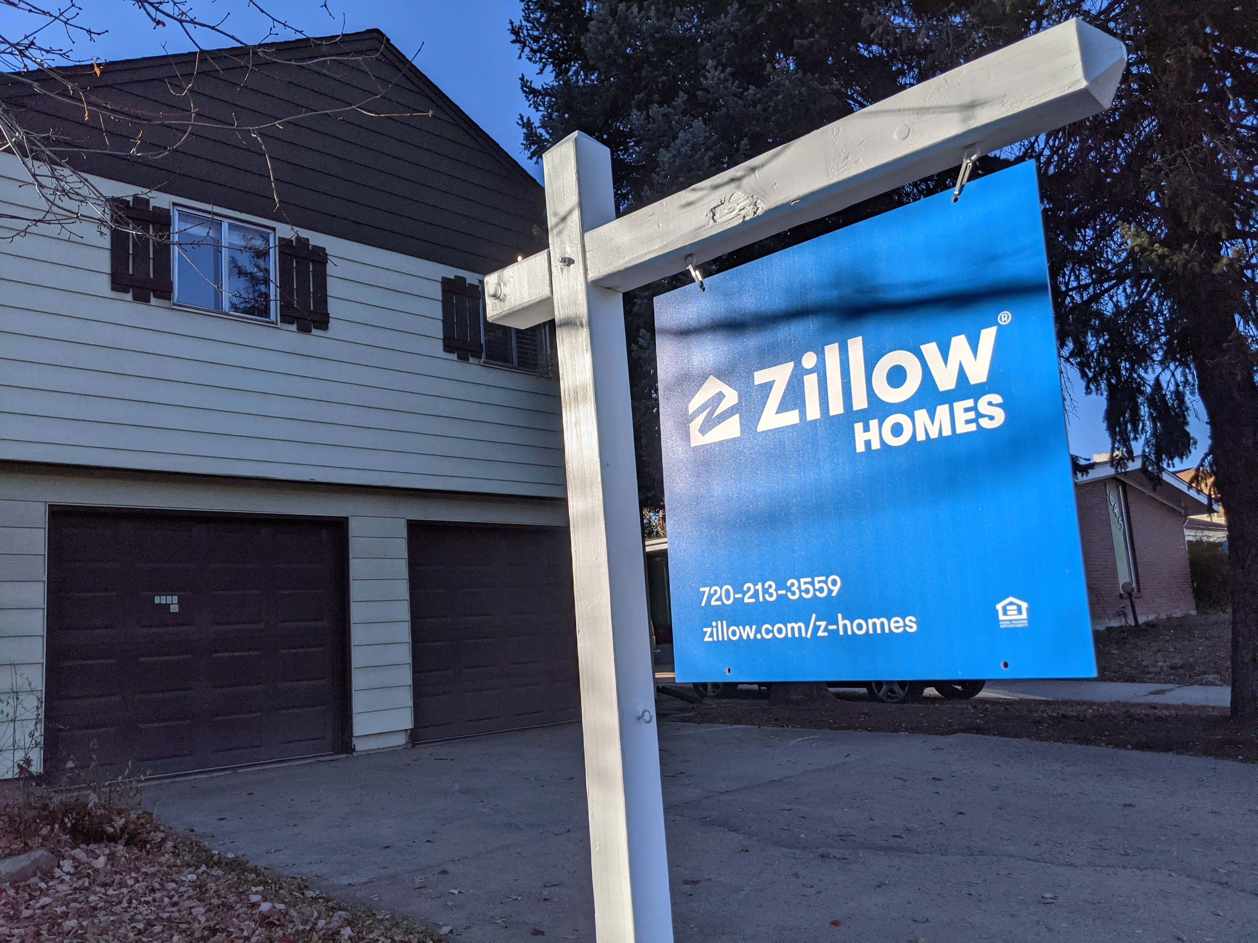 房屋 桌布 壁紙 — 一個亮藍色的Zillow Homes房地產標誌矗立在一棟有雙車庫的房子前，天空清澈湛藍。