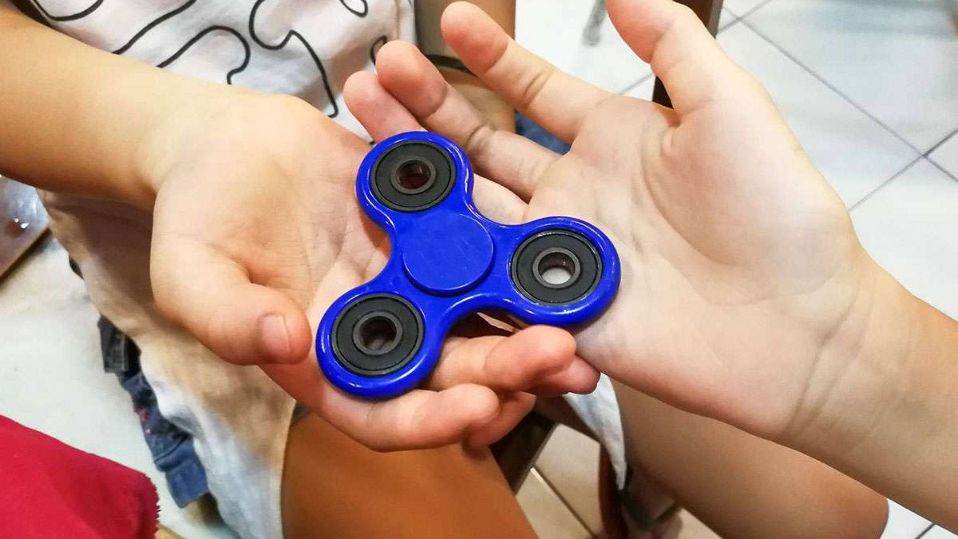 toys 桌布 壁紙 — 一個亮藍色的指尖陀螺輕輕地捧在合攏的雙手中，背景是瓷磚地板。