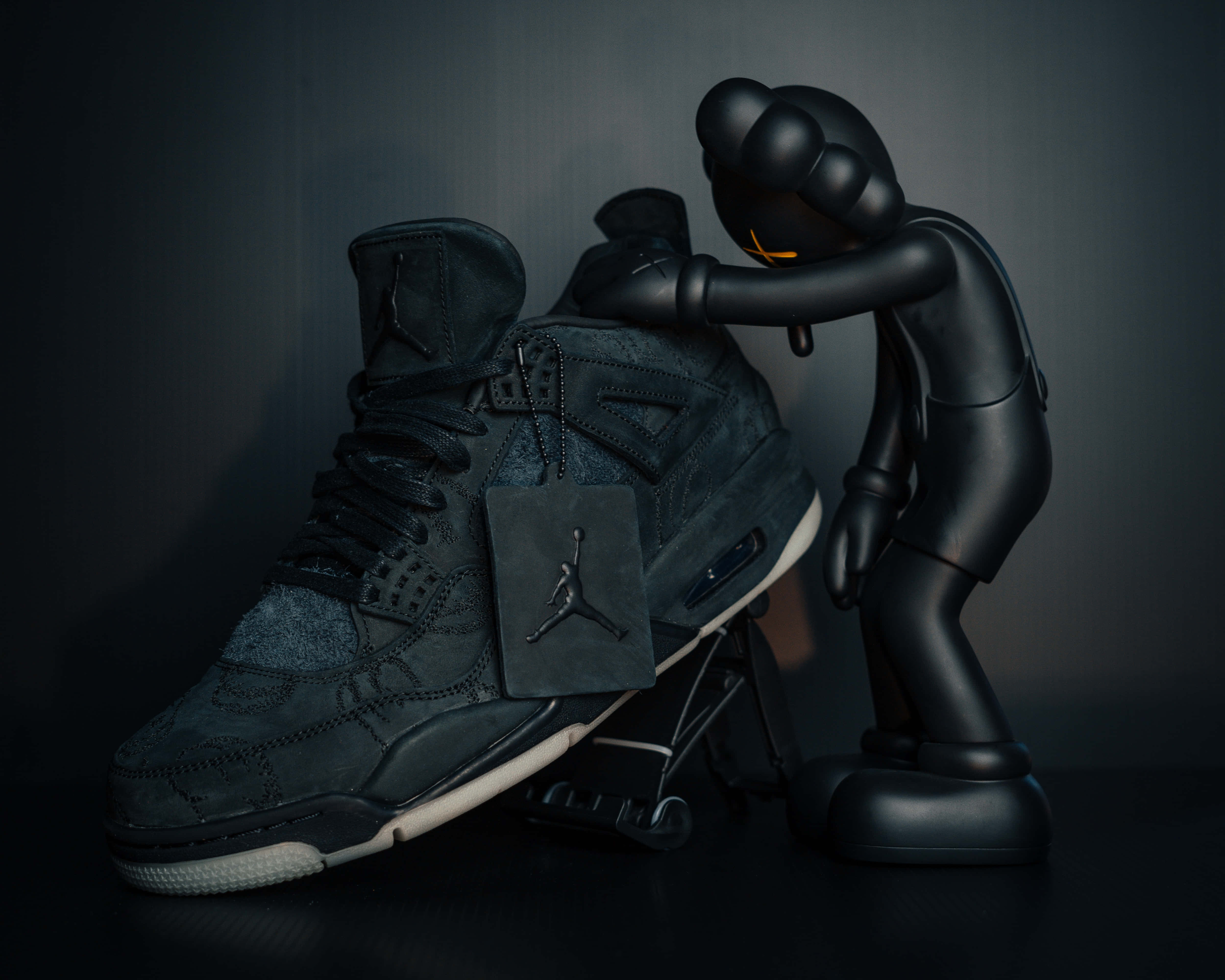 KAWS 桌布 壁紙 — 一個亮面黑色KAWS公仔在深色漸變背景前溫柔地擁抱一雙黑色Jordan運動鞋。
