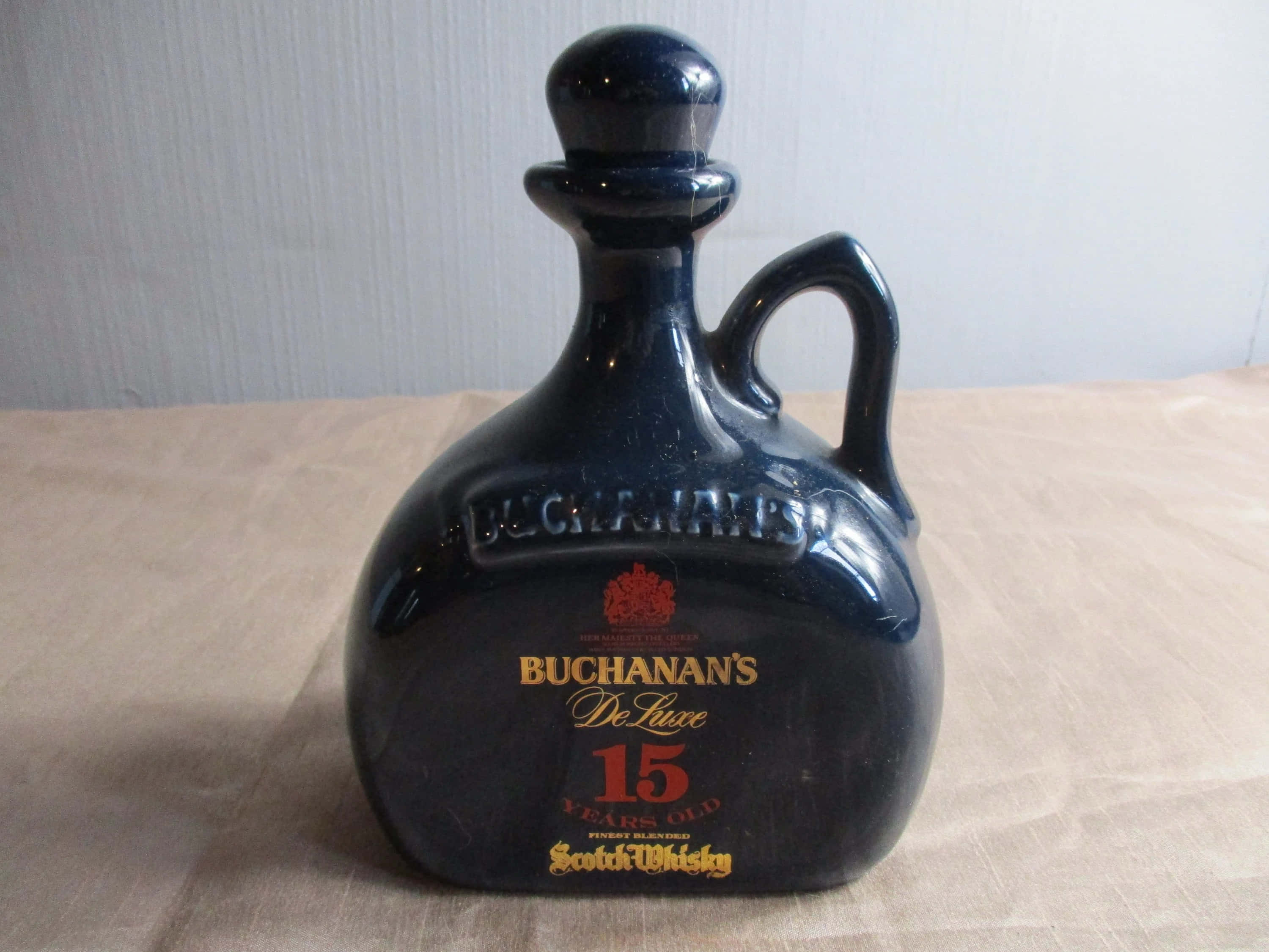 alcohol 桌布 壁紙 — 一個亮面黑色陶瓷壺裝的 Buchanan's 15 Year Old Scotch Whisky 放在米色布上，背景是淺灰色牆壁。