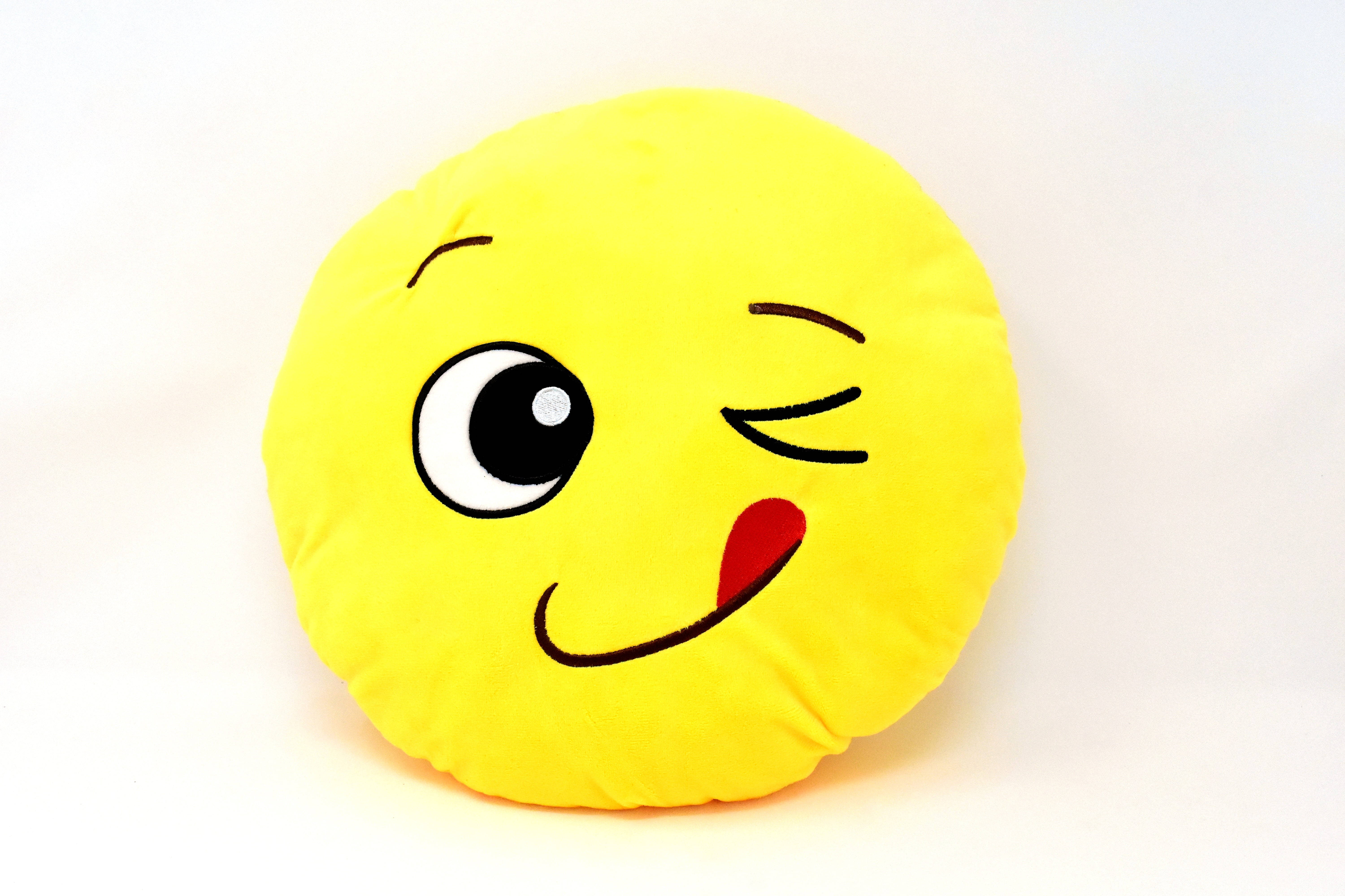 winky face 桌布 壁紙 — 一個亮黃色毛絨枕頭，形狀像一個眨眼的表情符號，紅色舌頭伸出，背景為白色。