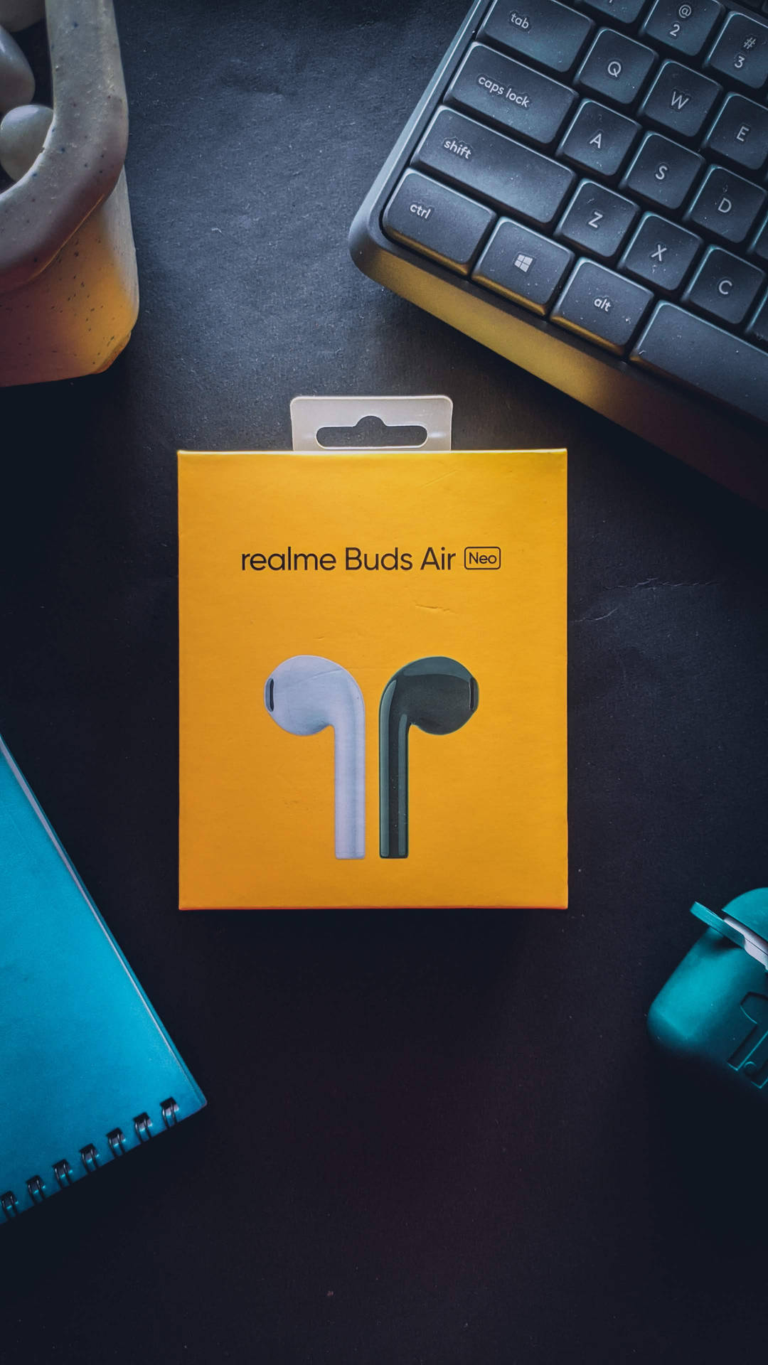 深色書桌上放著 realme Buds Air Neo 耳機盒，旁邊有鍵盤和筆記本。