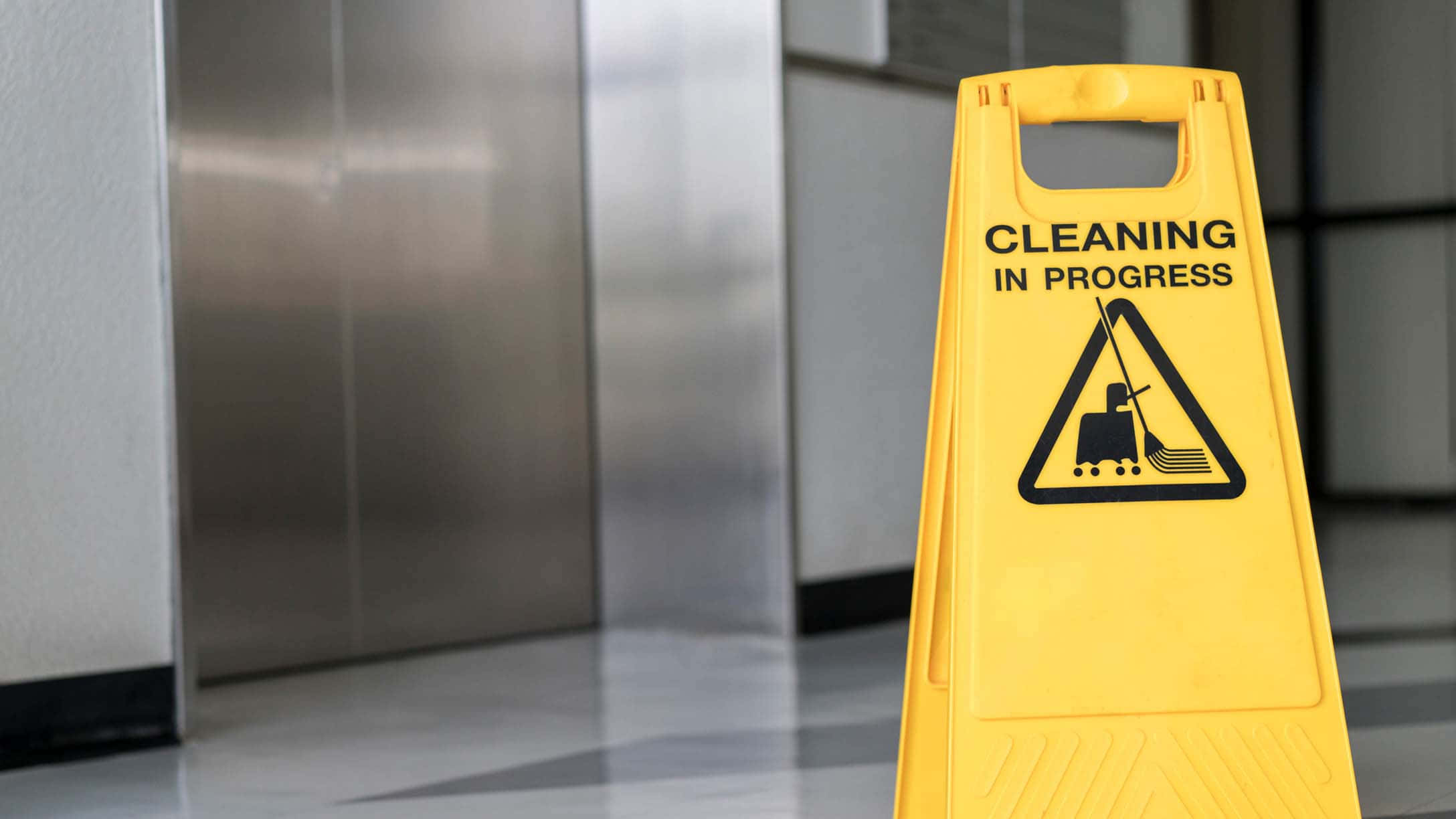 Cleaning 桌布 壁紙 — 一個亮黃色的「清潔進行中」警告標誌立在現代走廊中，靠近不鏽鋼電梯。