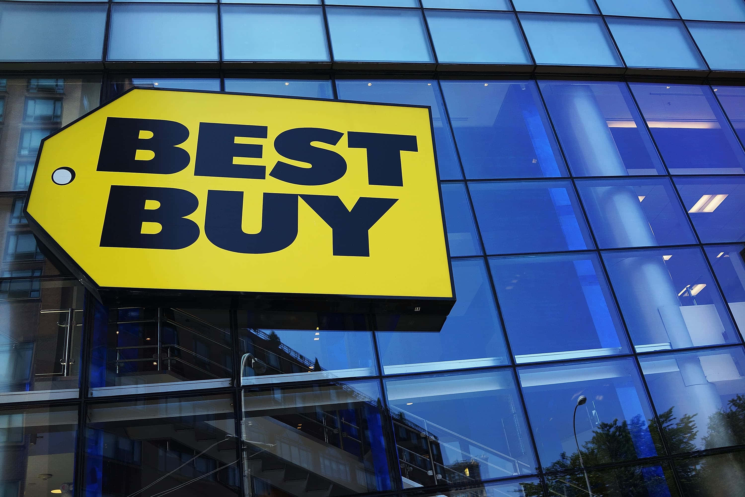 Best Buy 桌布 壁紙 — 一個亮黃色的Best Buy標誌醒目地掛在現代玻璃建築上，反射出藍天和城市環境。