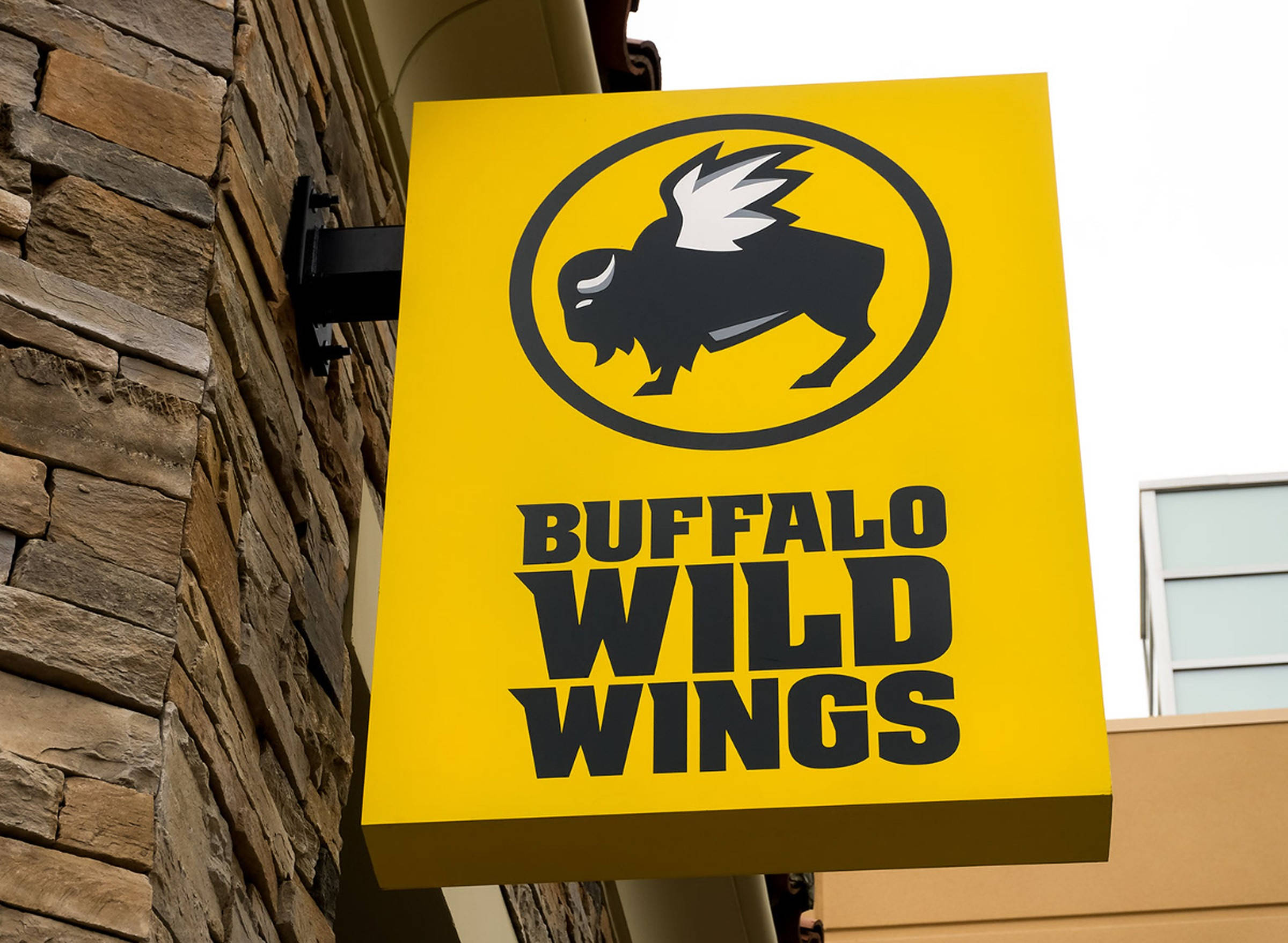 Wild 桌布 壁紙 — 一個亮黃色的Buffalo Wild Wings招牌，帶有黑色水牛標誌，懸掛在石頭建築外牆上。