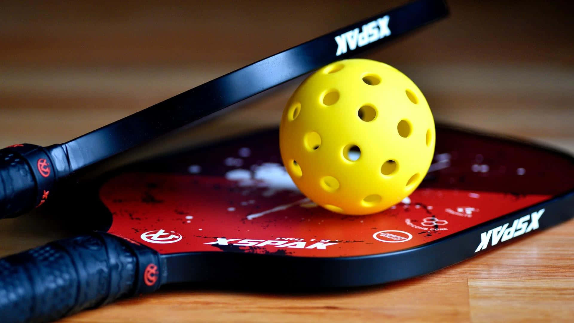 Pickleball 桌布 壁紙 — 一個亮黃色的匹克球放在一塊紅黑相間的球拍上，旁邊另一塊球拍斜靠在木地板上。