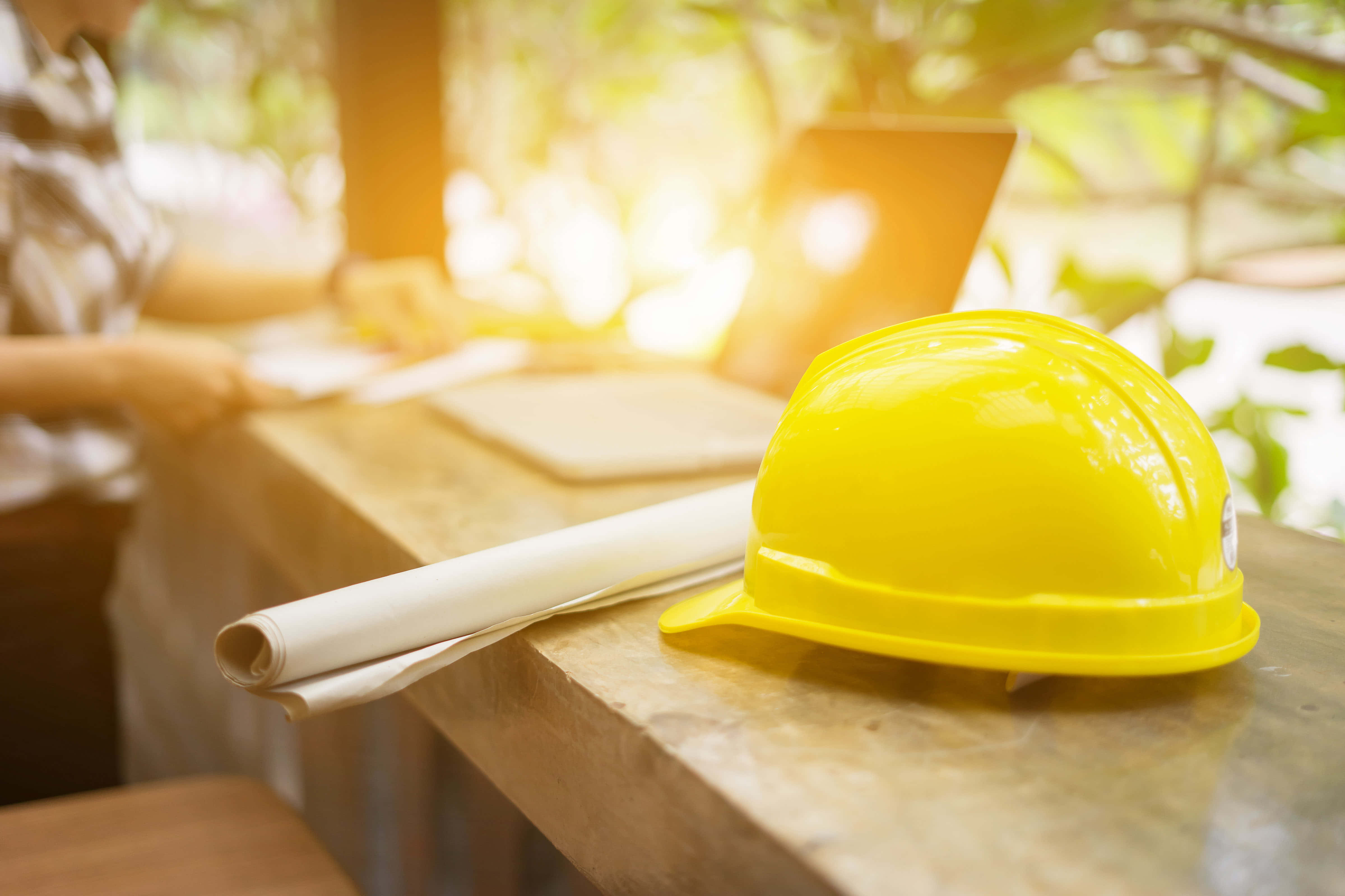 Engineer 桌布 壁紙 — 一個亮黃色的安全帽和捲起的藍圖放在木製工作檯上，一個人在陽光普照的戶外工作空間使用筆記型電腦。