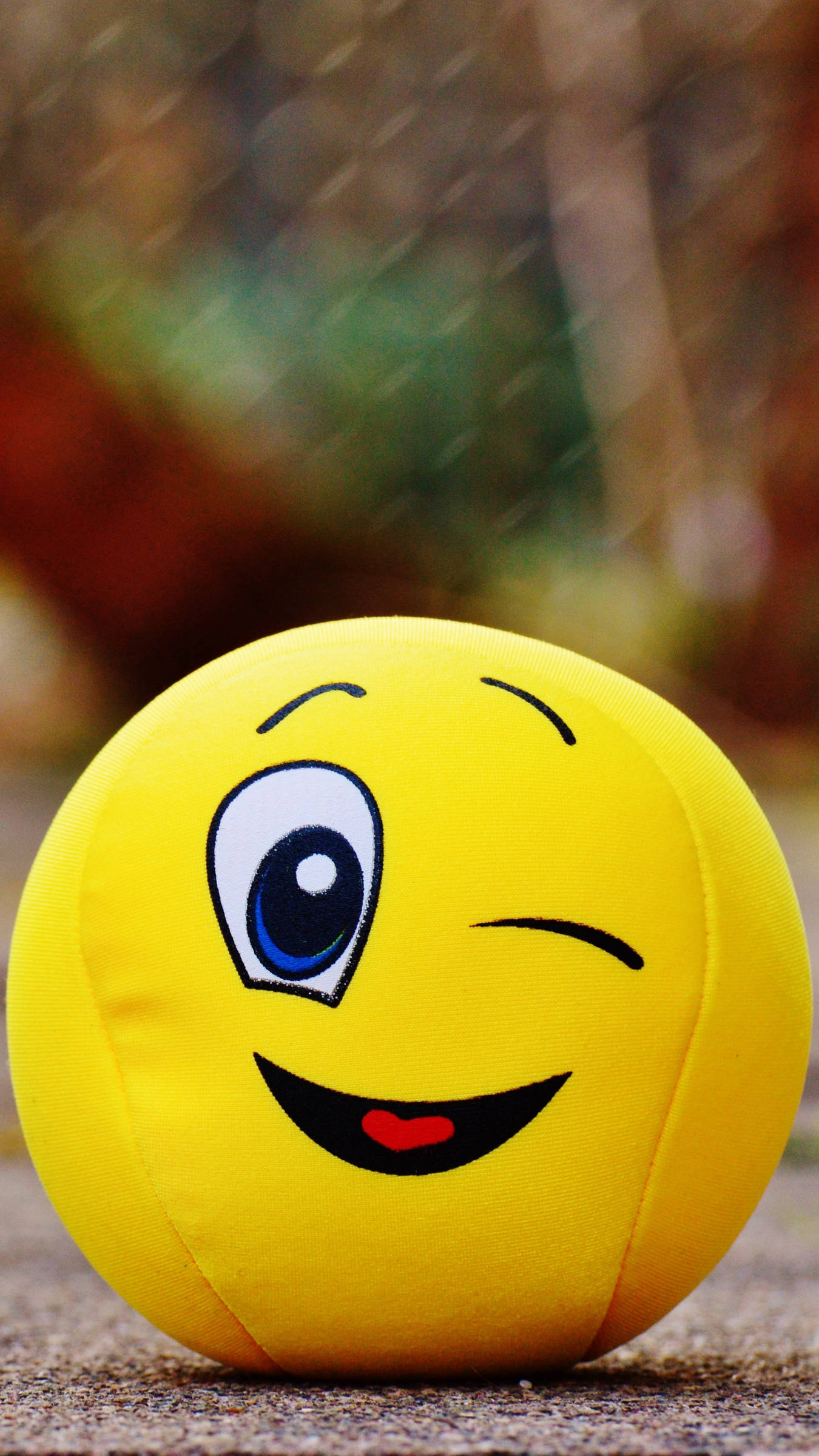 Smile 桌布 壁紙 — 一個亮黃色的毛絨表情符號球，一隻眼睜開，一隻眼閉上，正在眨眼，坐在有紋理的地面上，背景是模糊的鐵絲網圍欄。