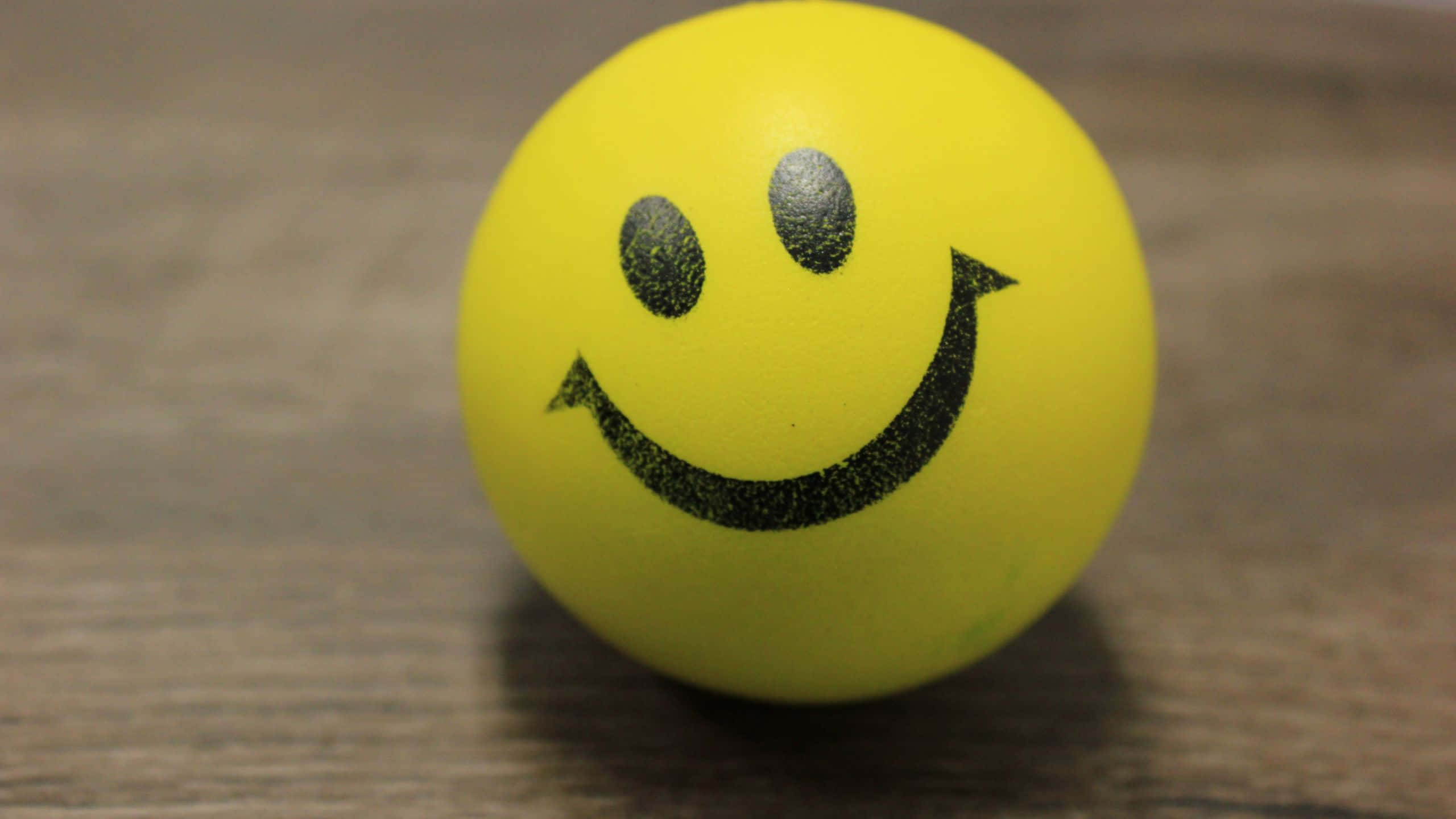 Smile Emoji 桌布 壁紙 — 一個亮黃色的泡棉球，上面有一個簡單的黑色笑臉，放在木質表面上。