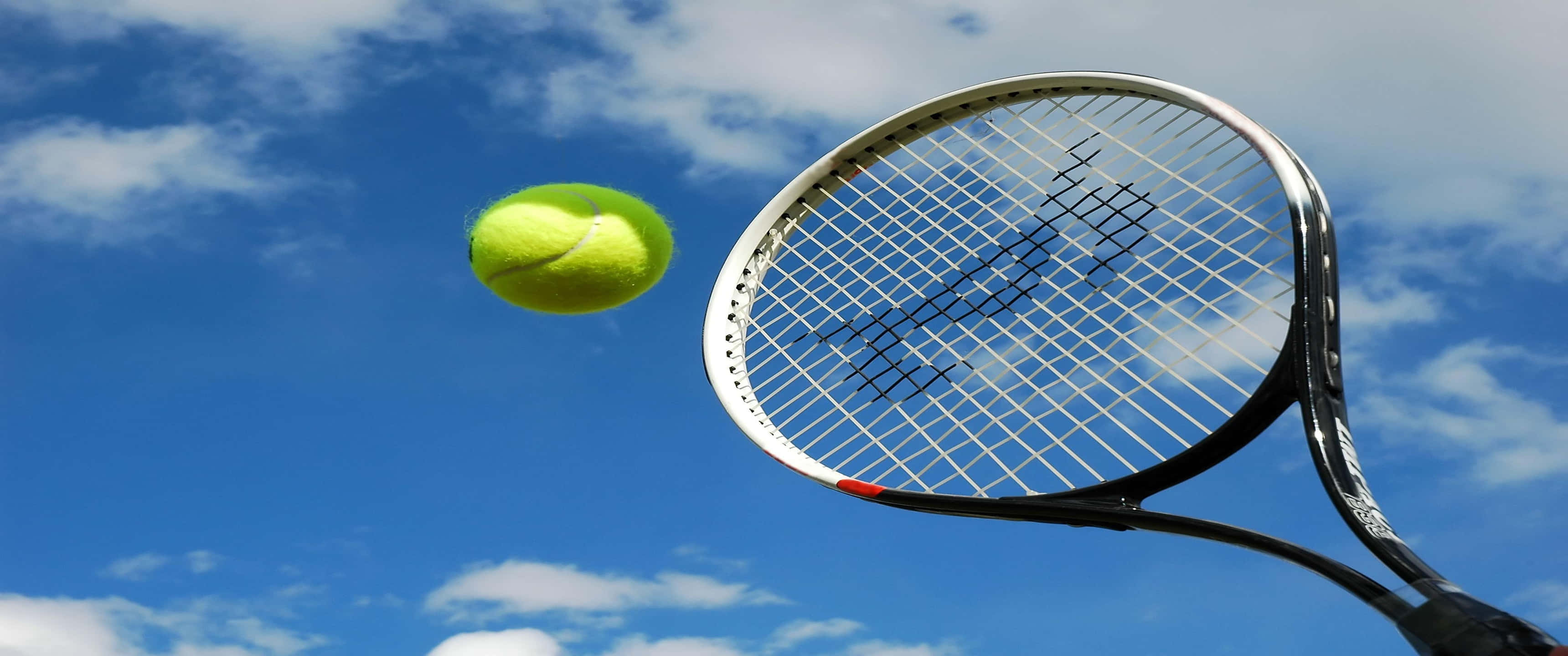 3440x1440p Tennis 桌布 壁紙 — 一個亮黃色的網球在鮮豔的藍天中懸浮，網球拍向上傾斜準備擊球。