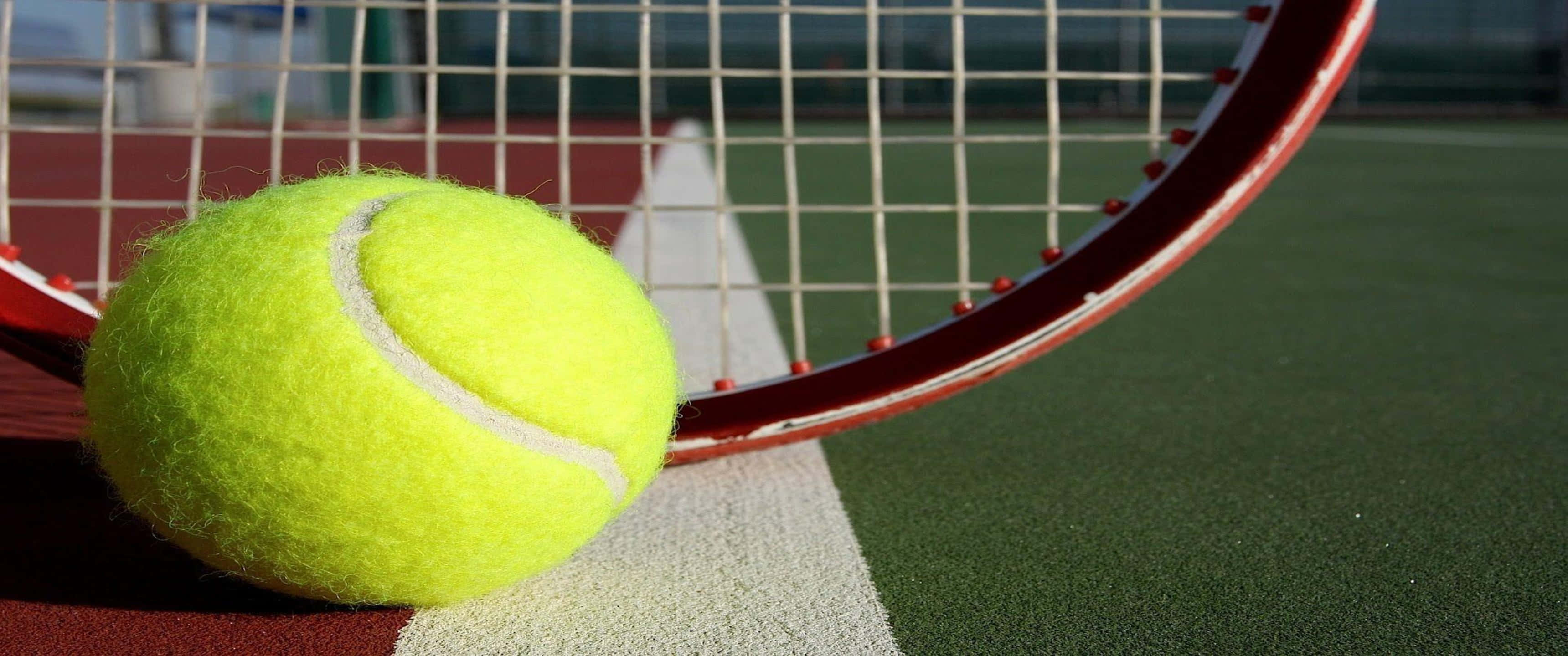 3440x1440p Tennis 桌布 壁紙 — 一個亮黃色的網球放在一個紅色球拍旁邊，球場是綠色和紅色，帶有白色線條。